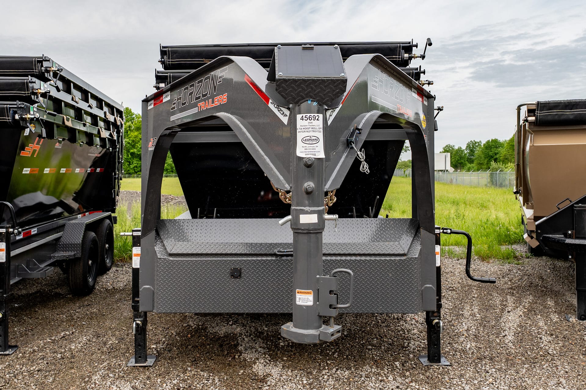 Rear view of a heavy-duty gooseneck trailer hitch and diamond-plate platform with other trailers in the background on gravel. WIth gray metal frame and warning labels visible.