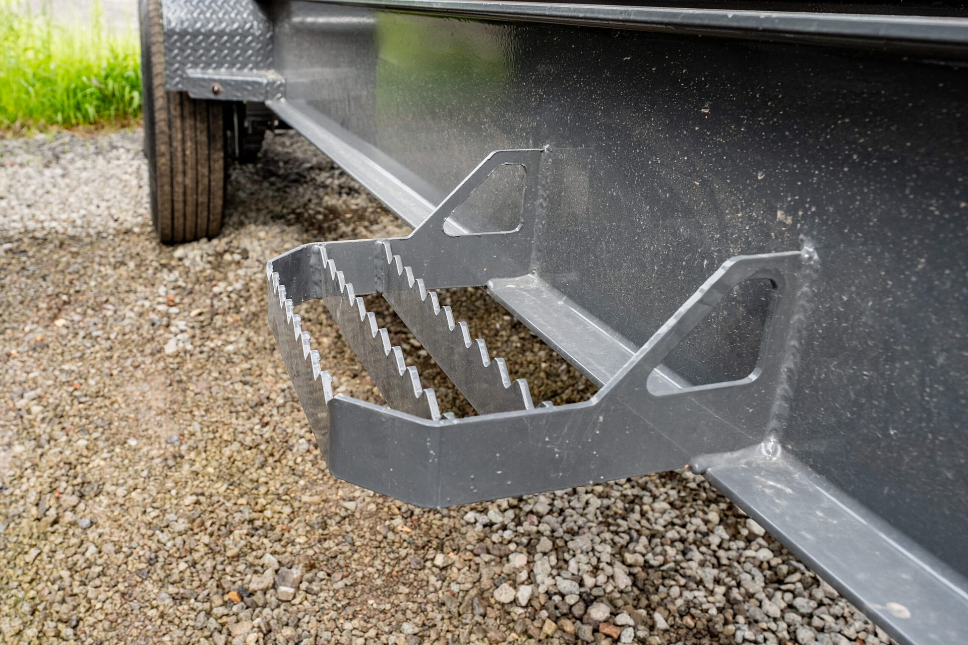 Close-up of metal stairs attached to a dark trailer, with a gravel ground beneath and a wheel in the background.
