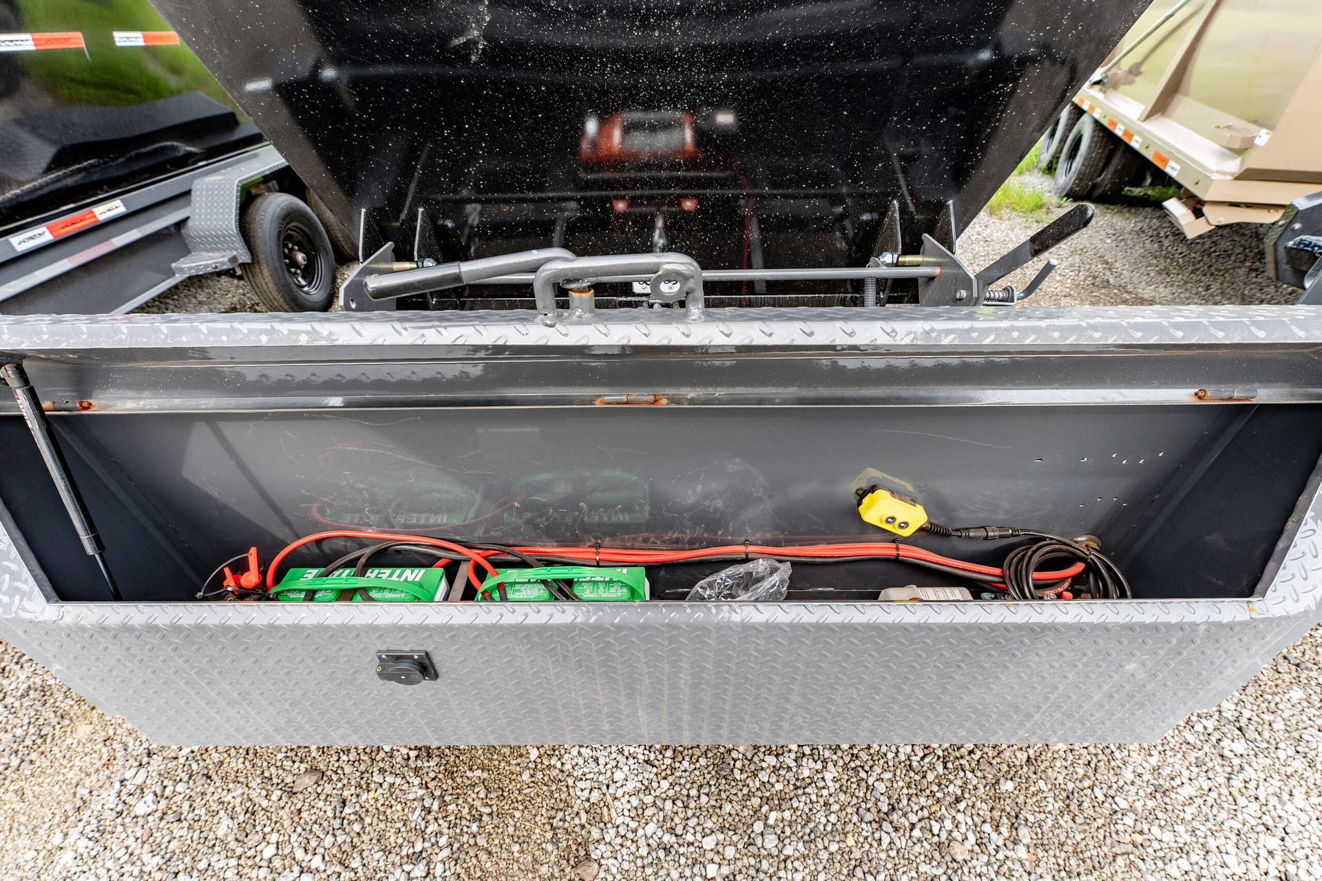Open trailer storage box with red and green battery packs, wires, and a hitch mechanism visible inside, on a diamond-plate metal floor above gravel ground