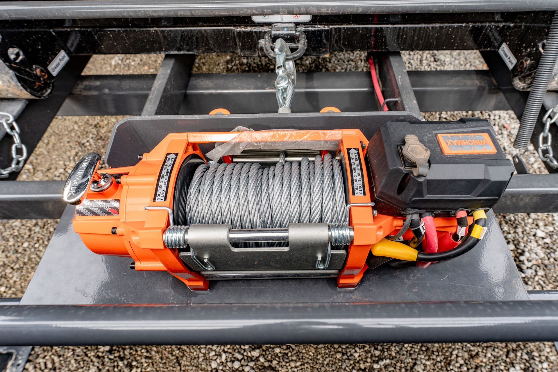 Close-up of an orange trailer winch with braided steel rope wound on the drum and a metal hook, mounted on a trailer frame.