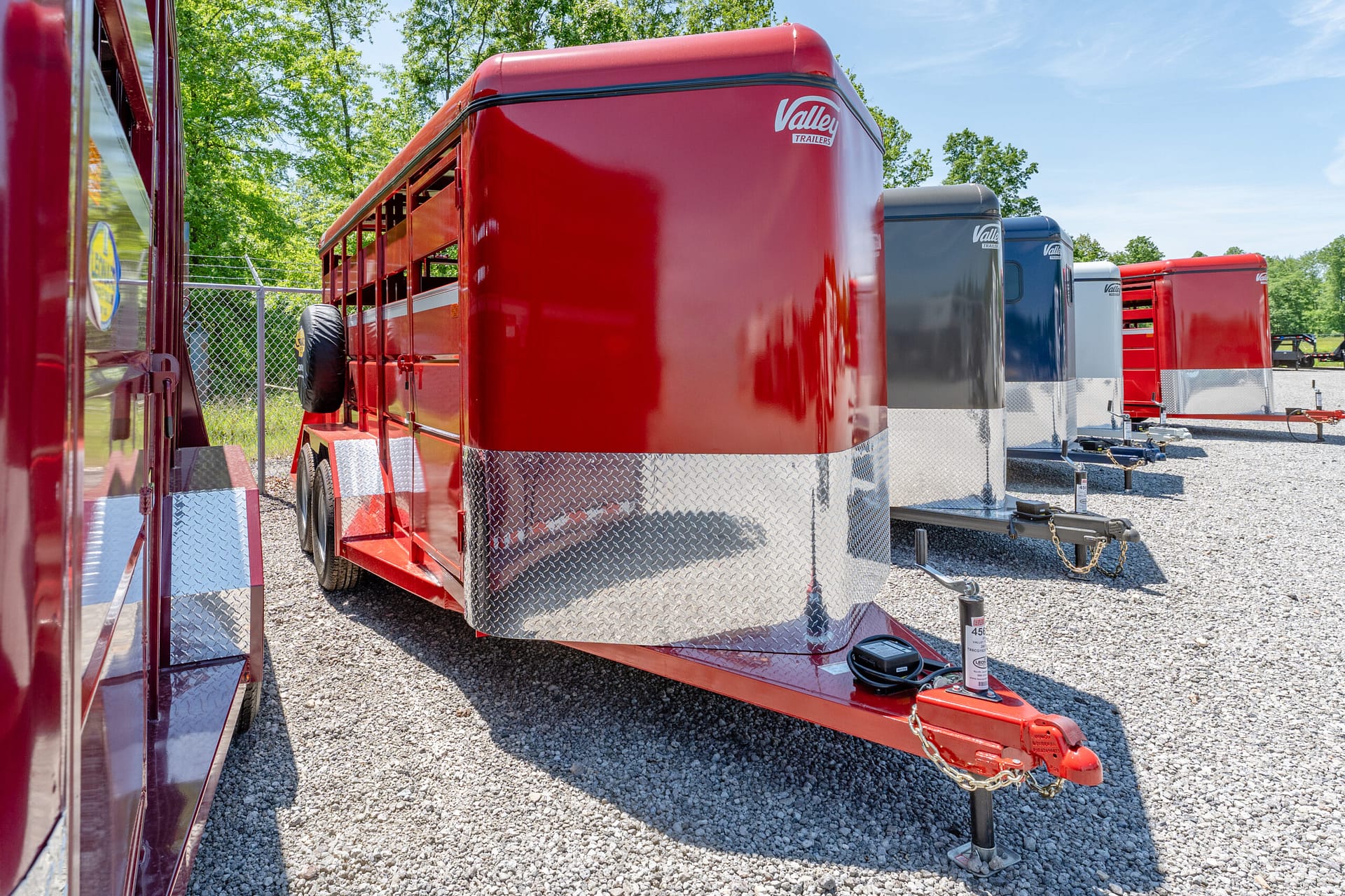 Row of colorful enclosed trailers parked on gravel, with trees in the background and a steel hitch in the foreground.