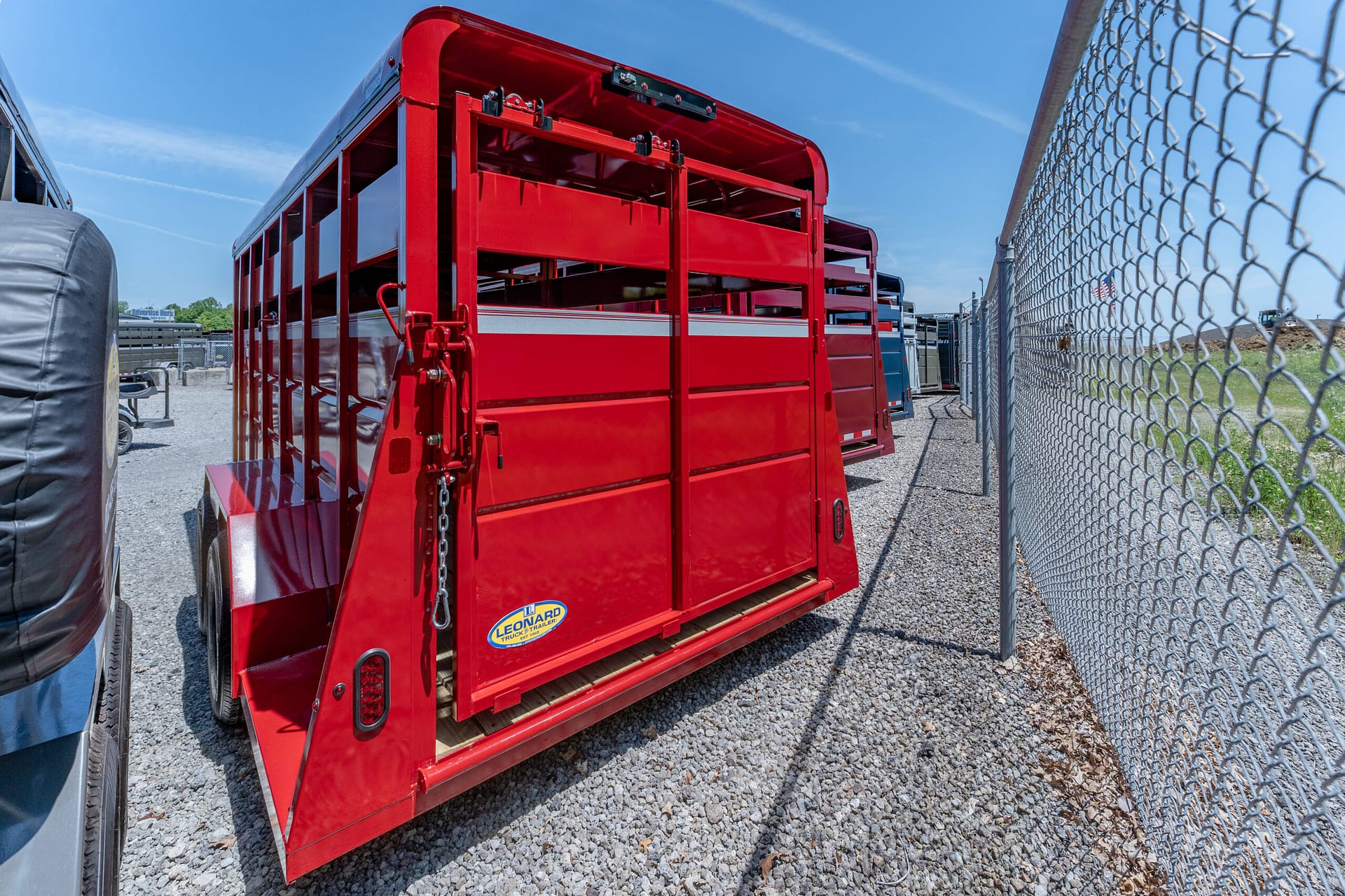 Red livestock/utility trailer with open side panels parked on a gravel lot beside a chain-link fence under a blue sky.