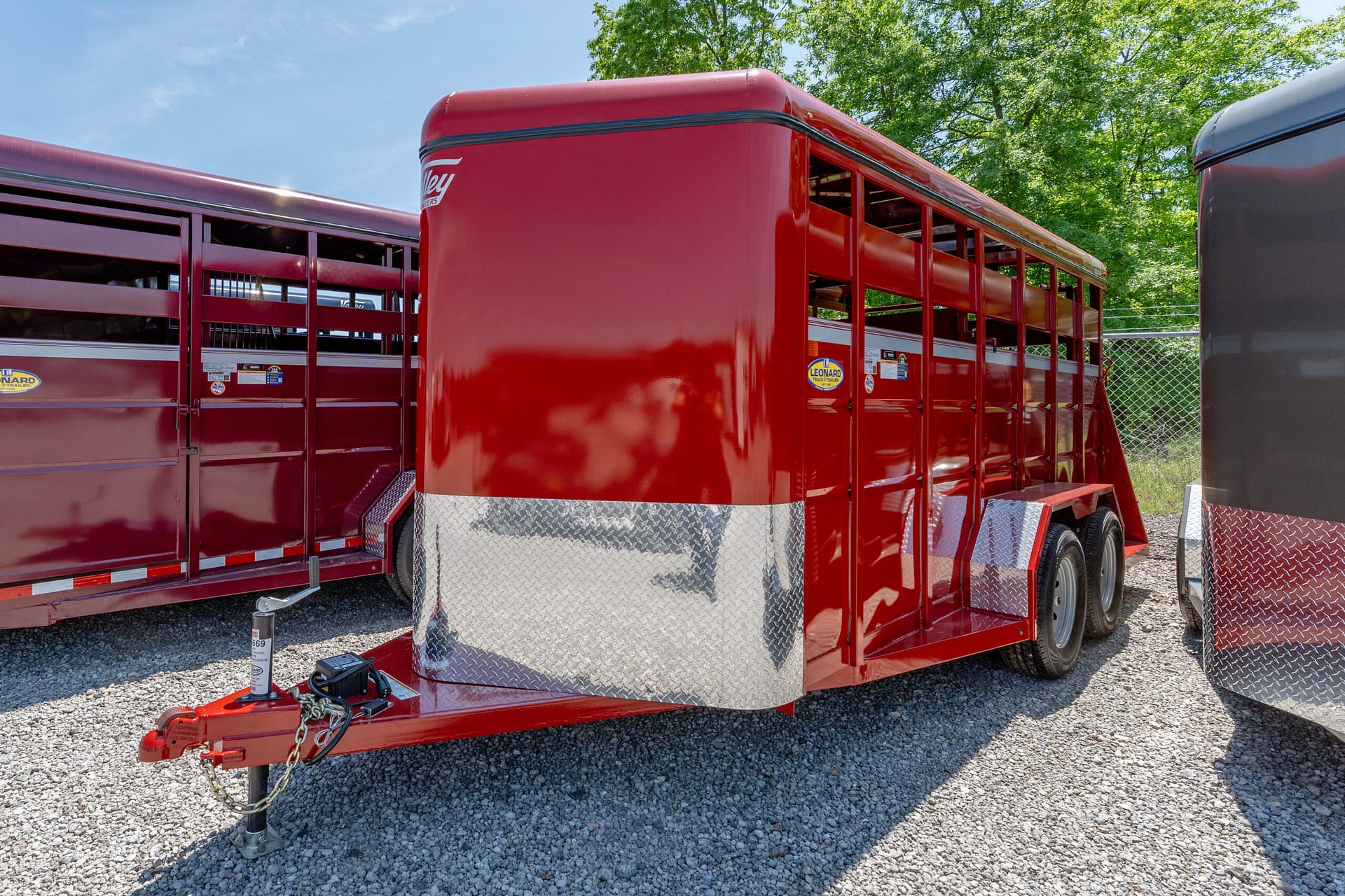 Red glossy trailer with diamond-plate lower edge and hitch on gravel, outdoors in bright daylight.