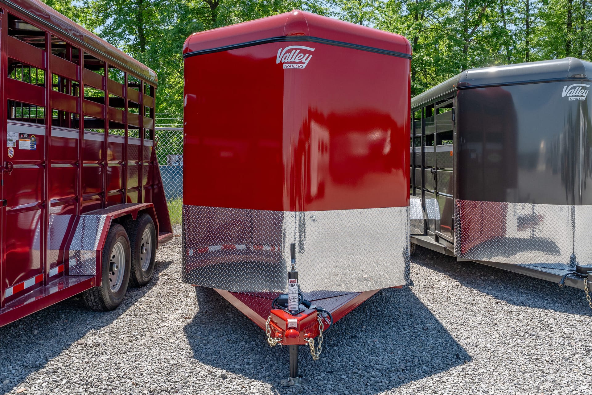 Red Valley Trailers horse trailer with diamond-plate skirting parked on gravel beside a gray trailer, trees in the background.