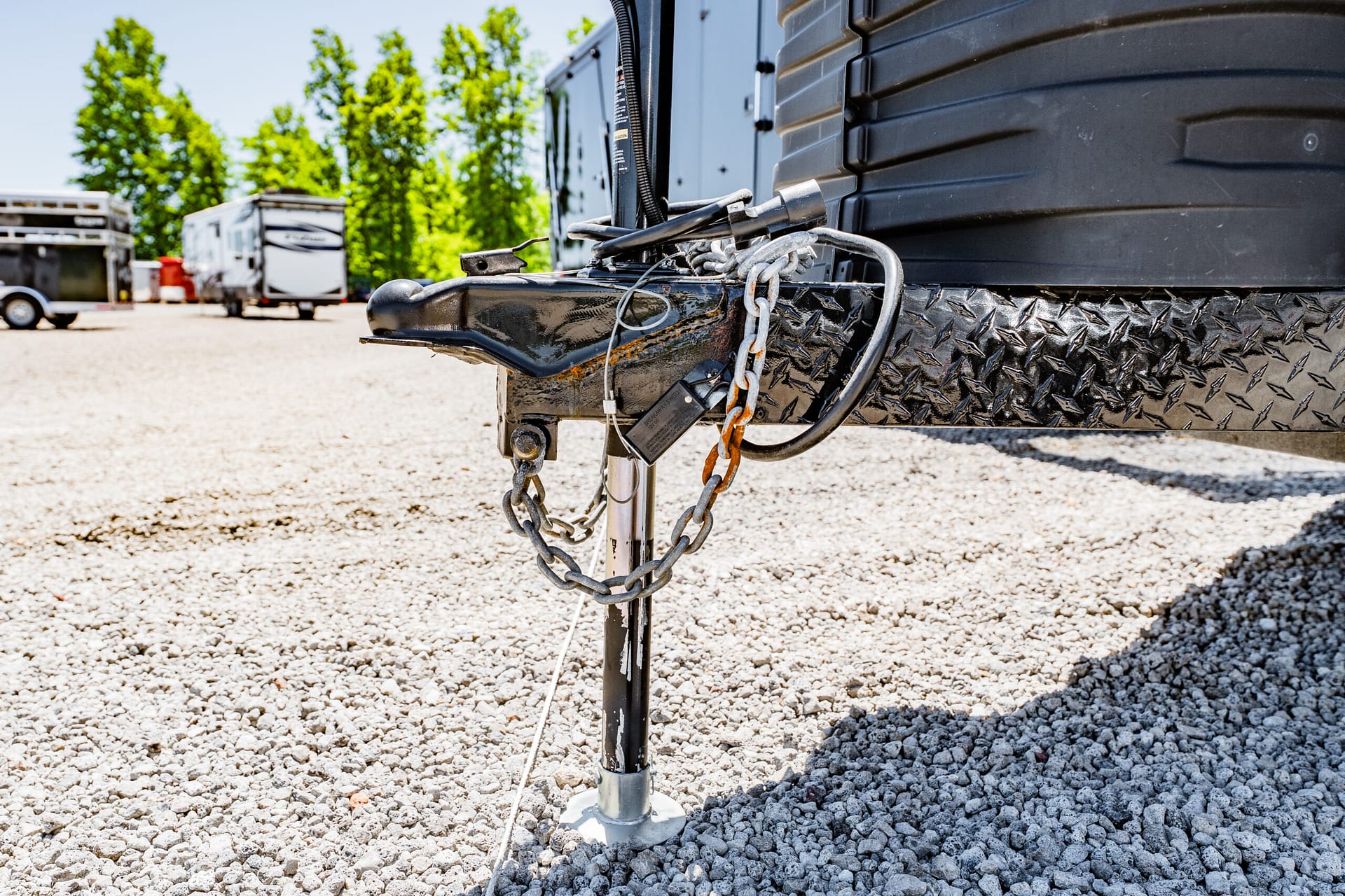 Close-up of a trailer hitch with safety chains and a lock on a gravel lot, RVs in the background.