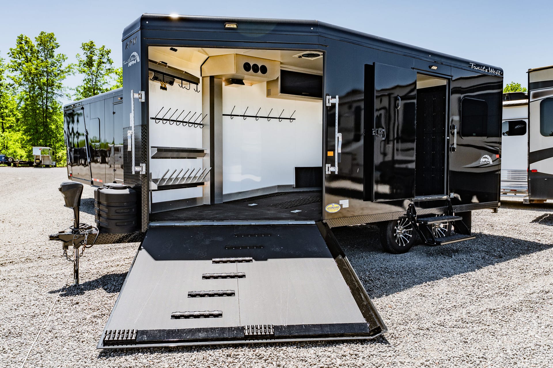 Black trailer with its side wall open, showing white interior, wall racks for gear, and a lowered metal ramp on a gravel lot