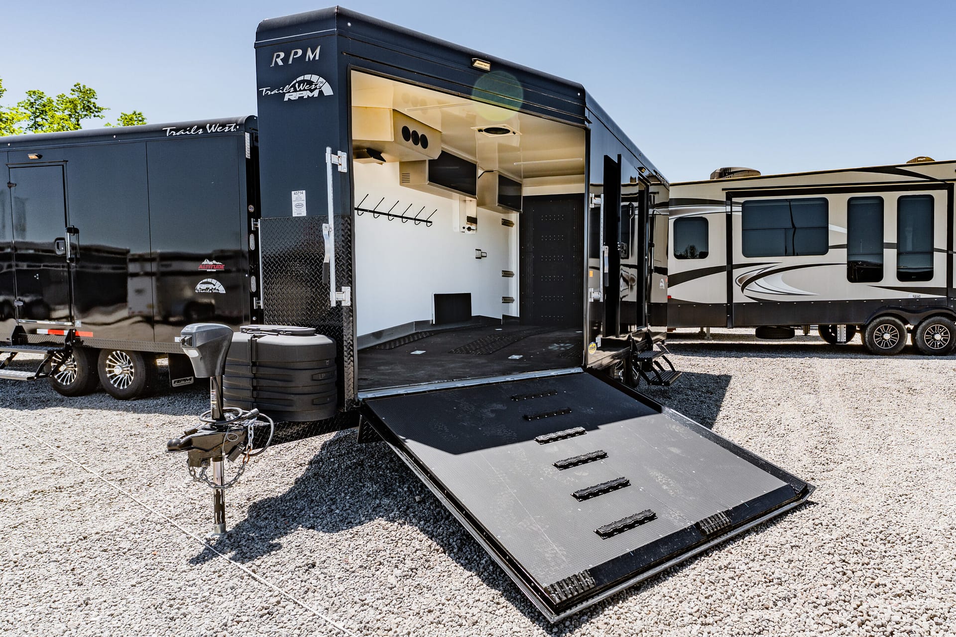 Black toy hauler trailer with an open rear ramp, exposing an interior storage/work area beside another RV in a gravel lot