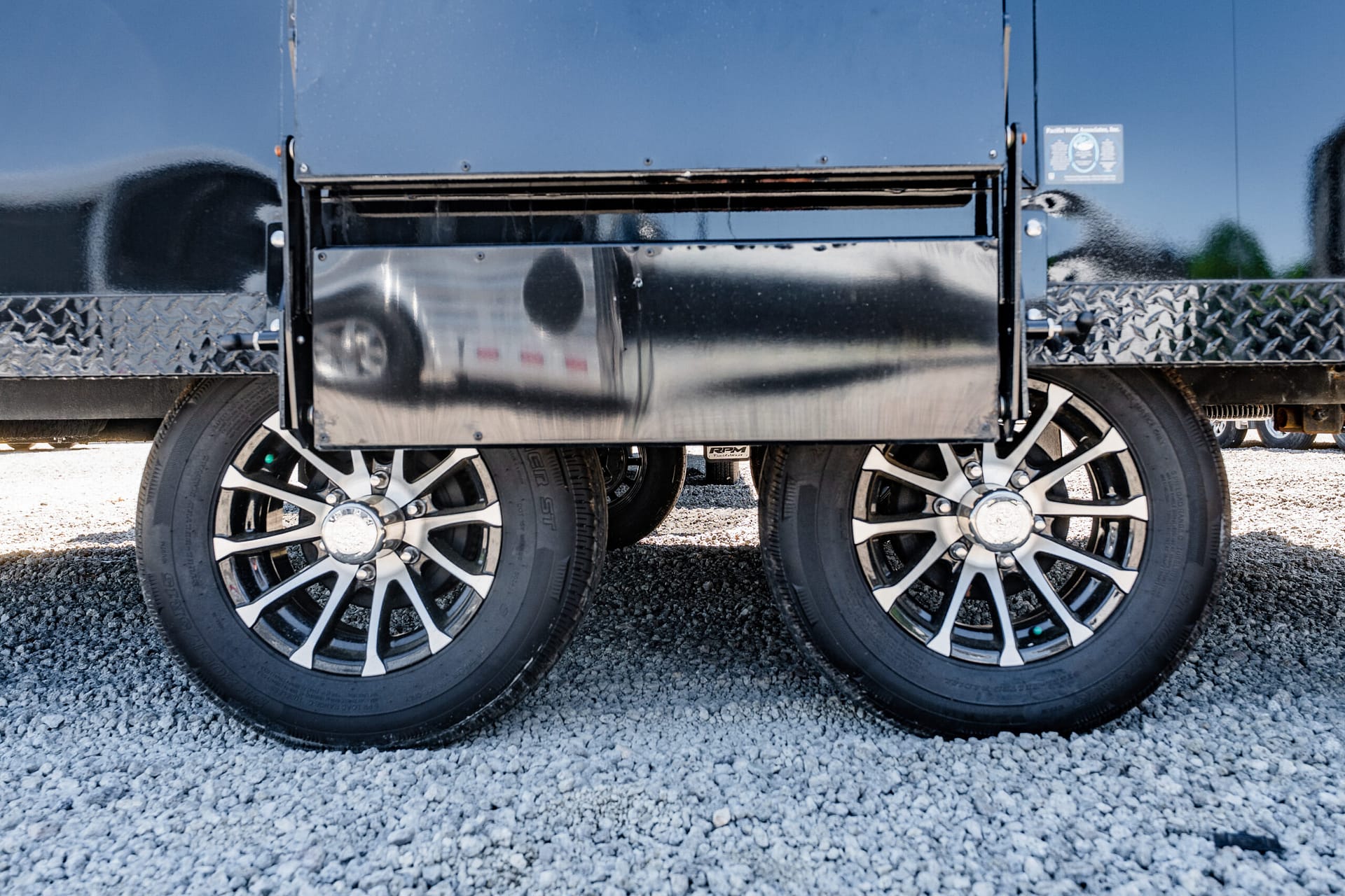 Close-up of dual trailer wheels with shiny chrome rims and diamond-plate side on gravel surface
