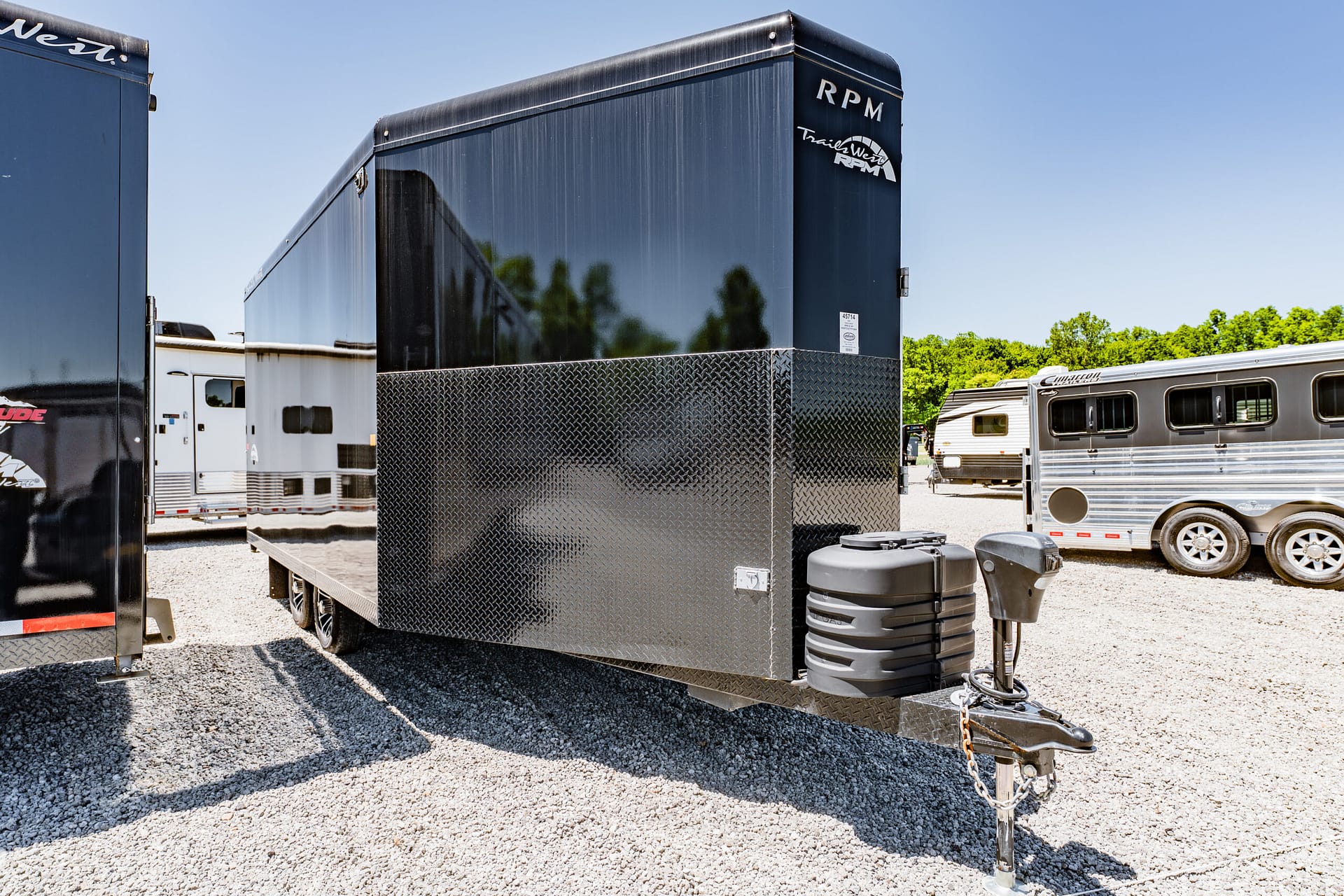Black enclosed trailer with diamond-plate lower half in a trailer lot, other trailers visible against a blue sky.