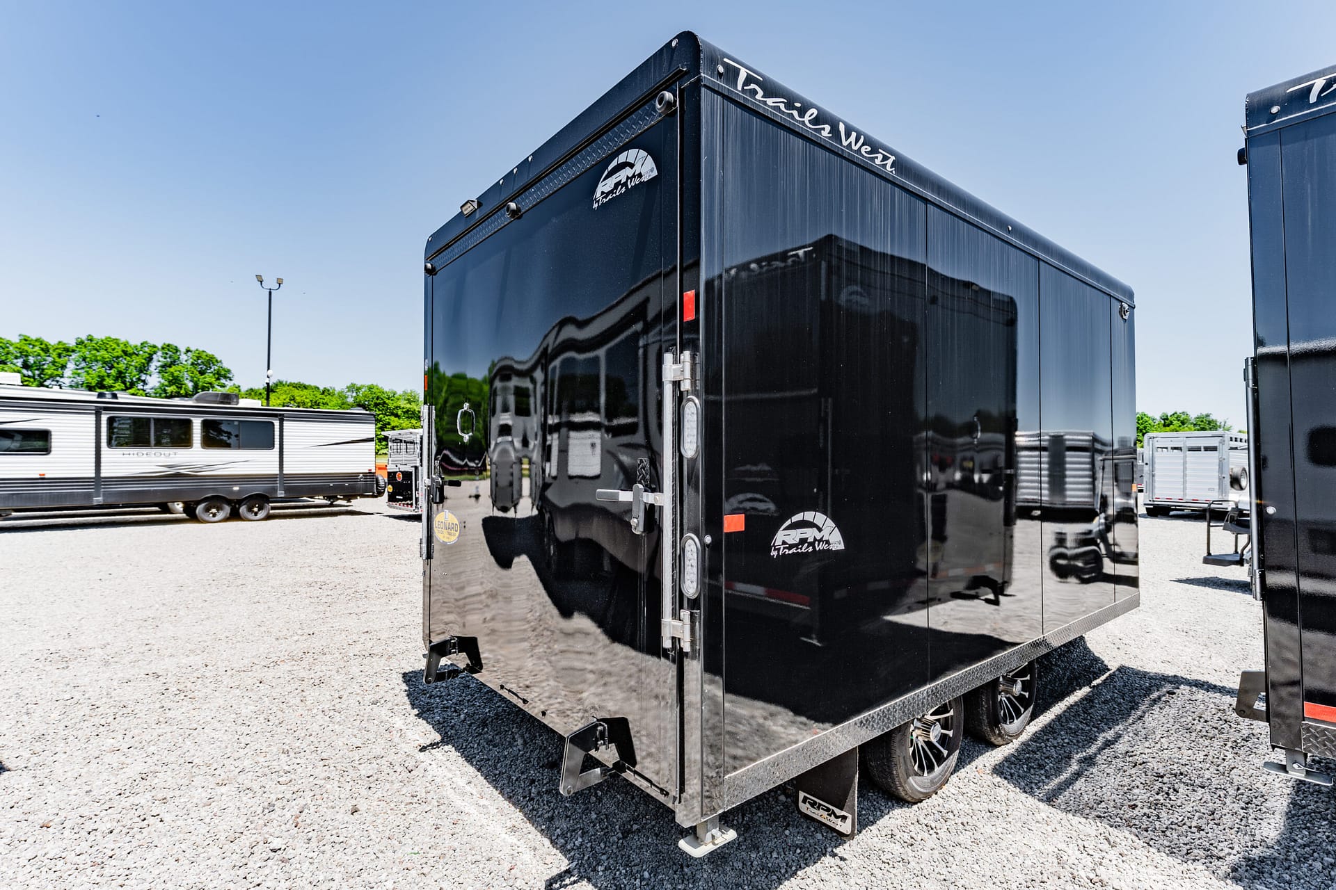 Black glossy enclosed trailer in a gravel lot, with reflections of other trailers on its side and a clear blue sky above.