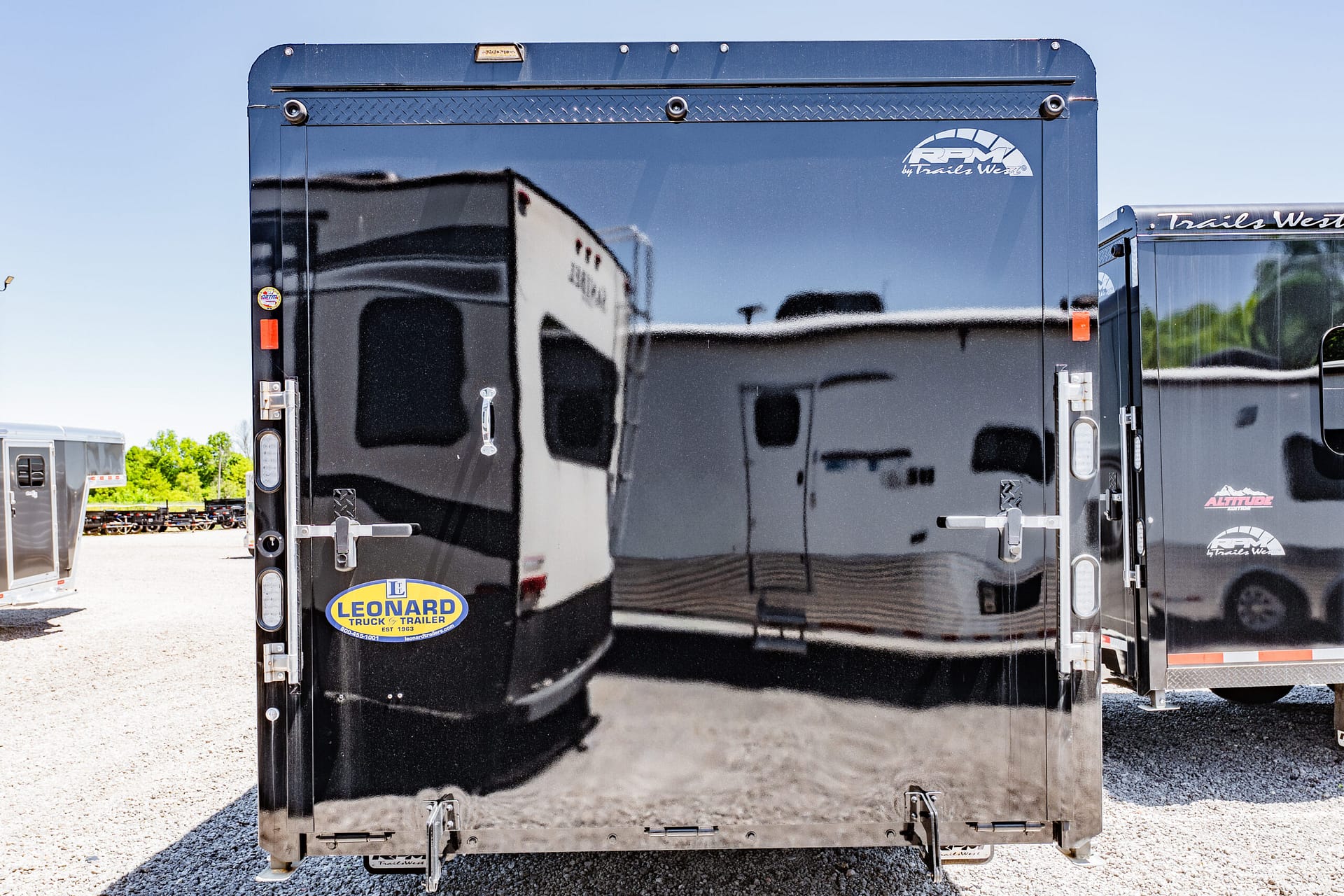 Rear view of a glossy black trailer with brand logos (RPM by Trails West) and a Leonard sticker, in an outdoor lot with other trailers nearby