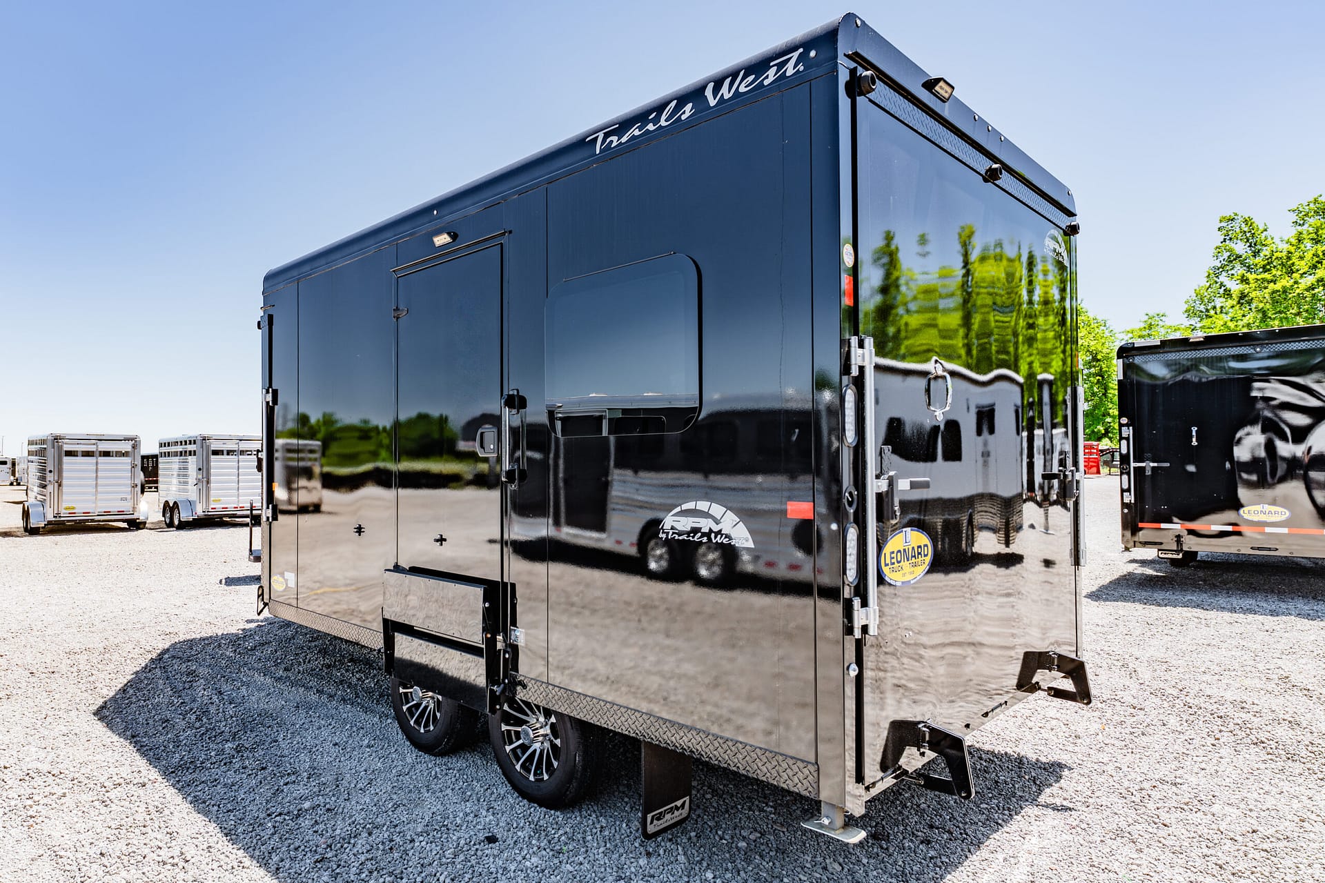 Black glossy trailer with 'Trails West' branding in a gravel lot, reflections visible on the side panels