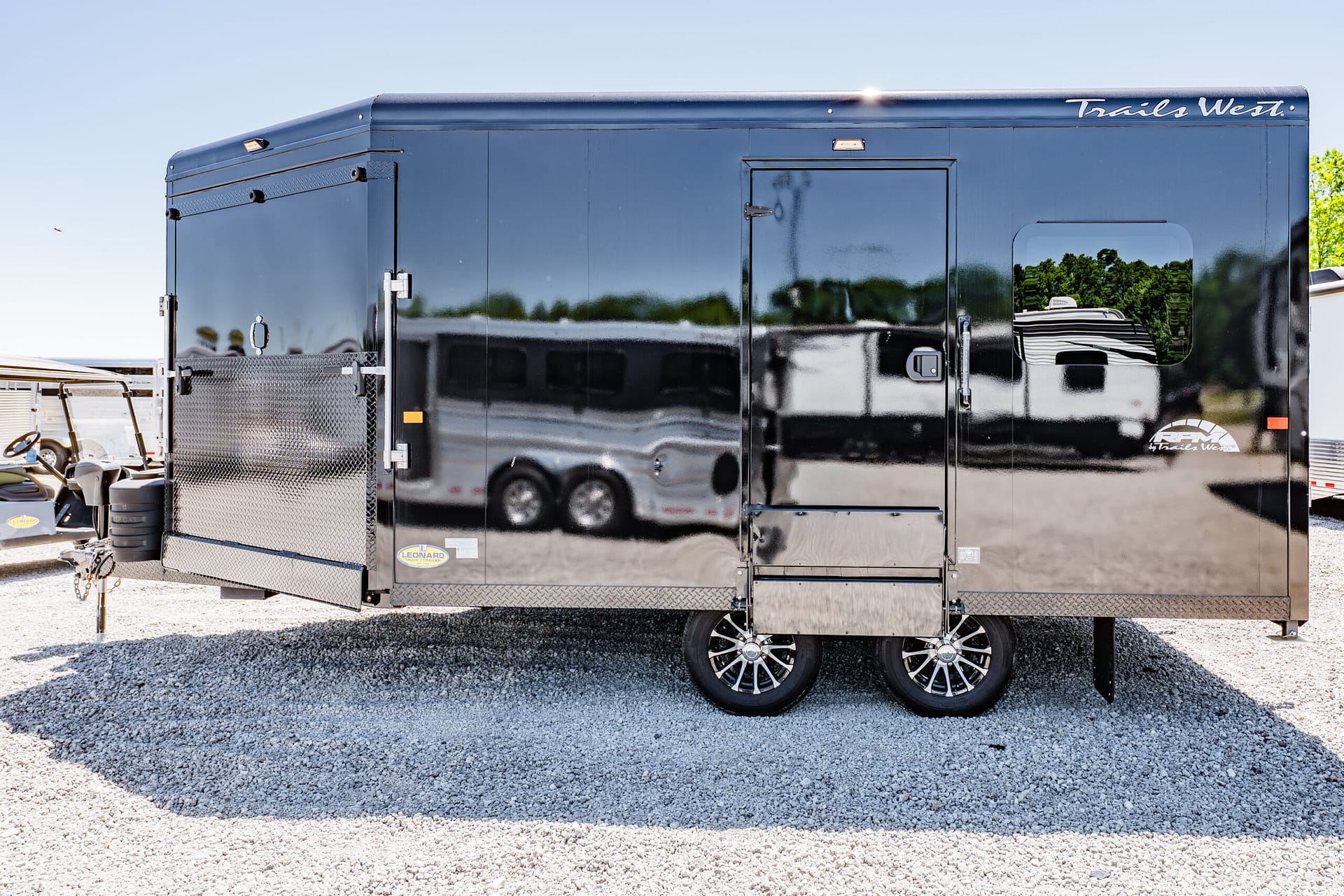 Black Trails West enclosed trailer with a side door and window, parked on a gravel lot; its glossy surface reflects another trailer nearby.