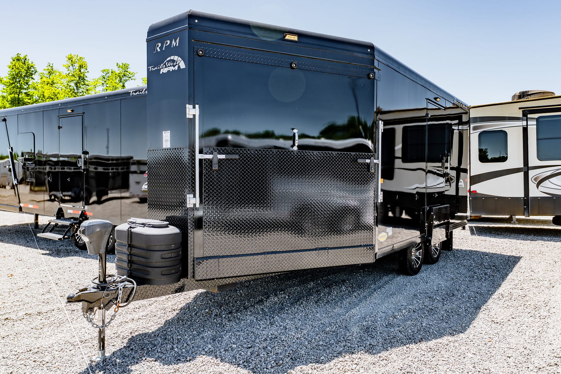 Blue travel trailer with diamond-plate front, hitch and propane tank in foreground on a gravel lot with other RVs nearby.