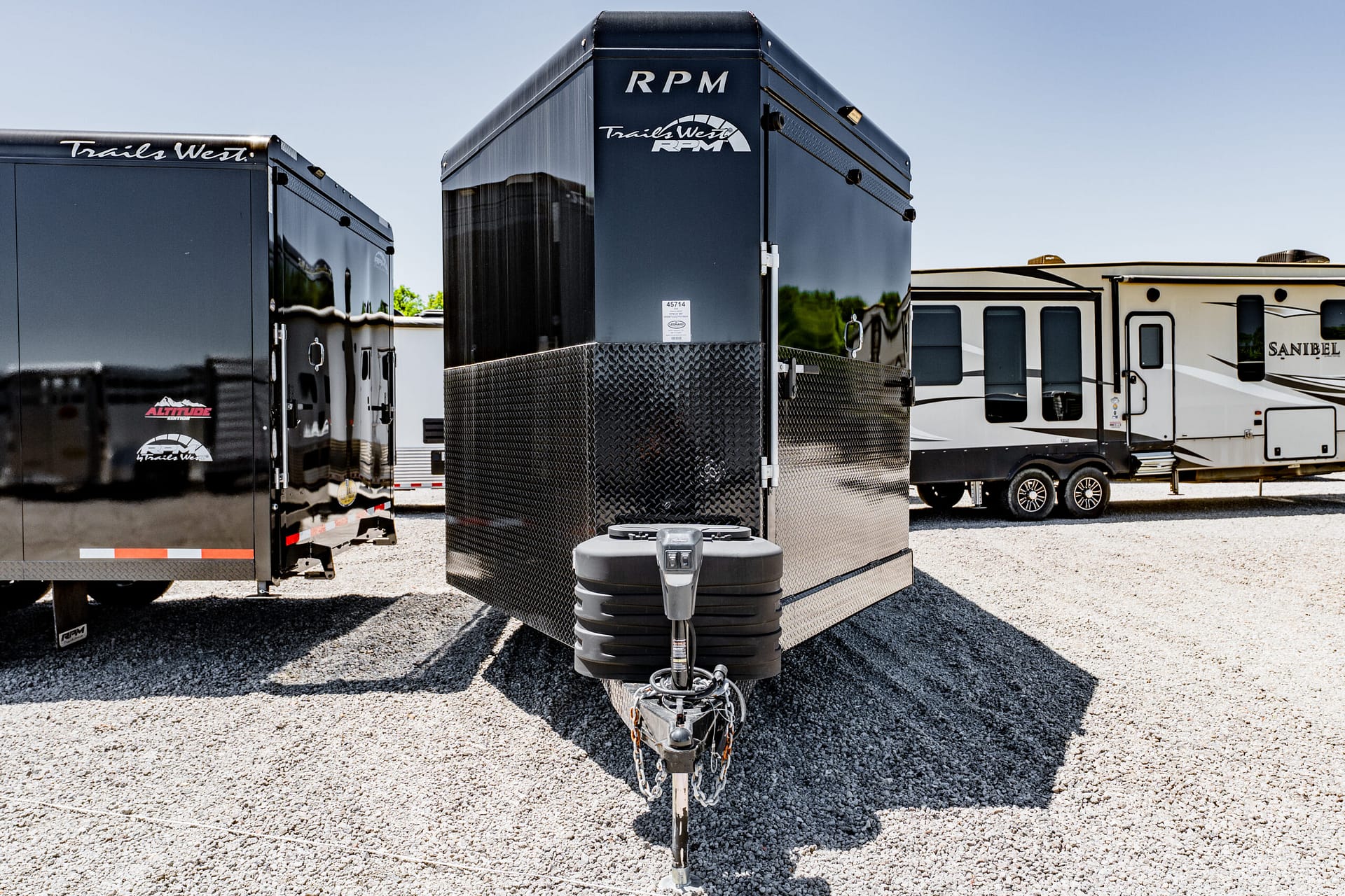 Front view of a black metal trailer with diamond-plate lower half, hitch in foreground, other trailers in background on gravel lot