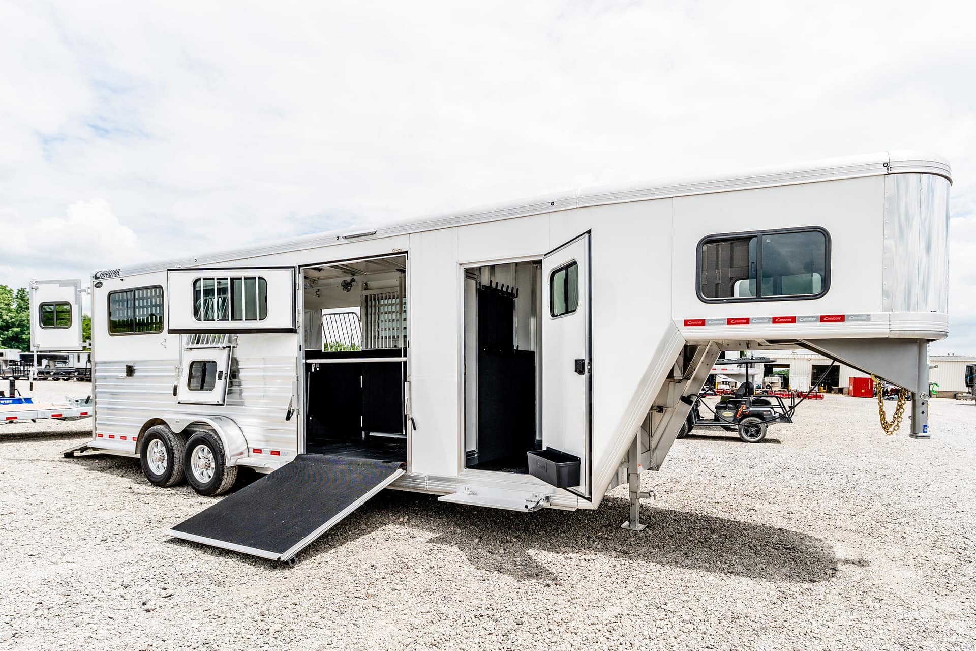 White horse trailer with side door open and a lowered ramp on a gravel lot, revealing interior stalls.