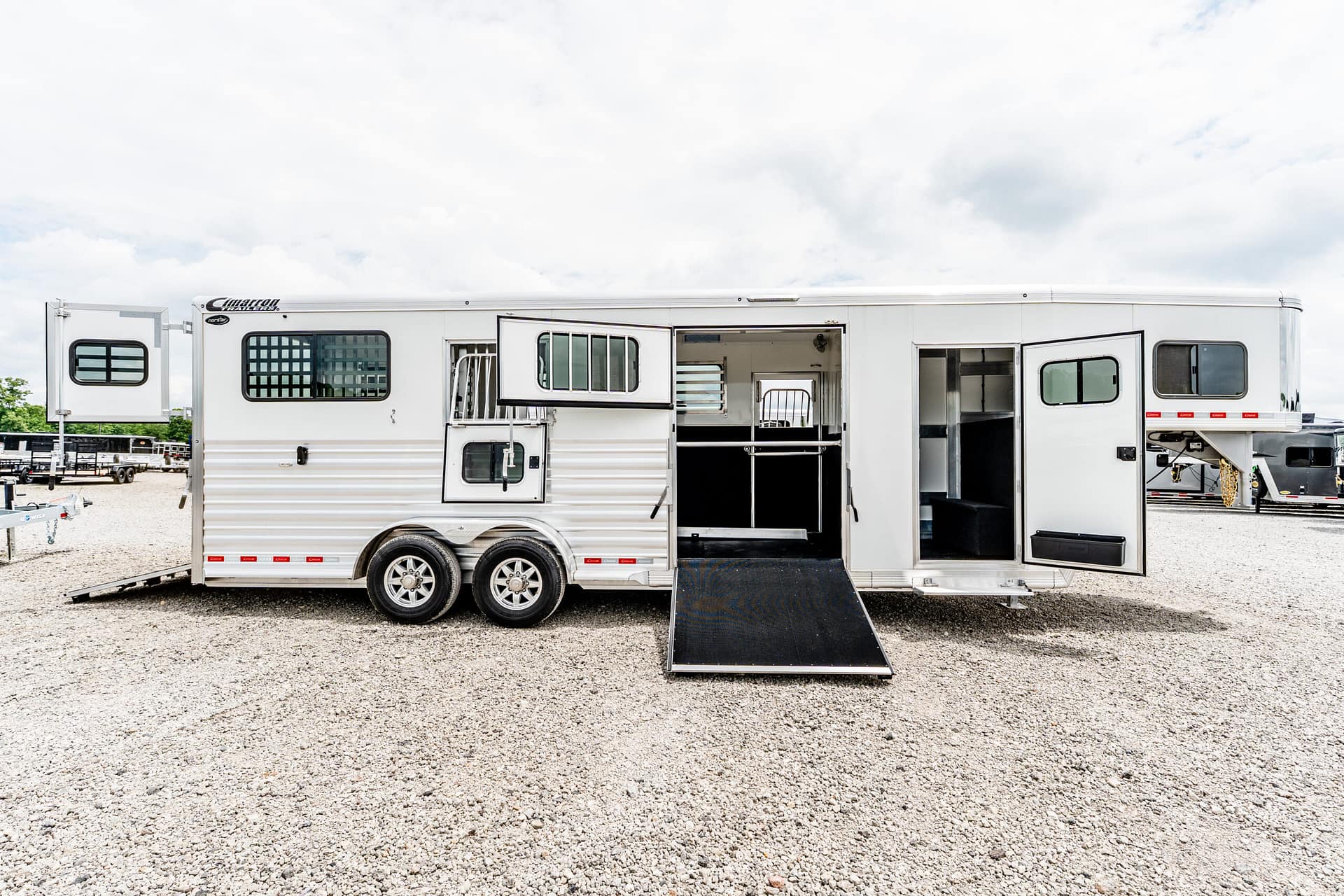 Silver horse trailer with side access doors open and a lowered ramp, parked on a gravel lot under a cloudy sky.