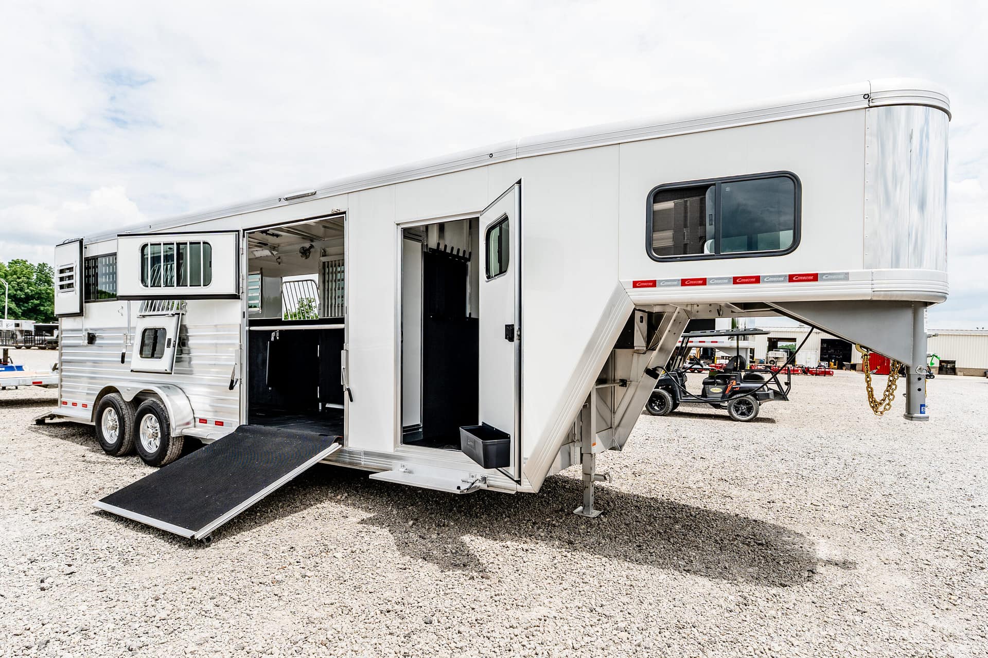 White horse trailer with its side doors open and a lowered ramp, parked on a gravel lot under a cloudy sky.
