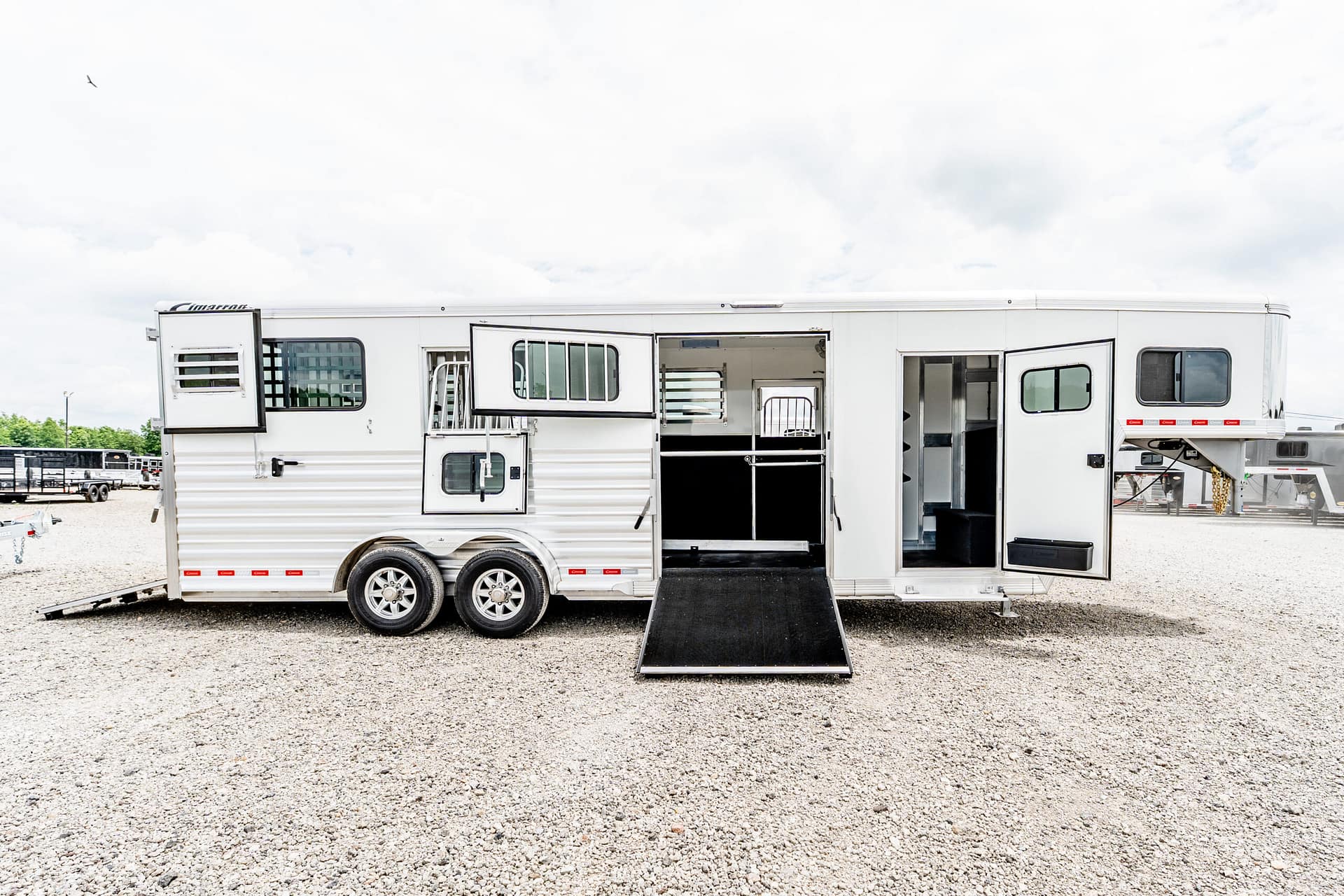 White horse trailer with open side entrance and lowered ramp on a gravel lot, showing stalls inside