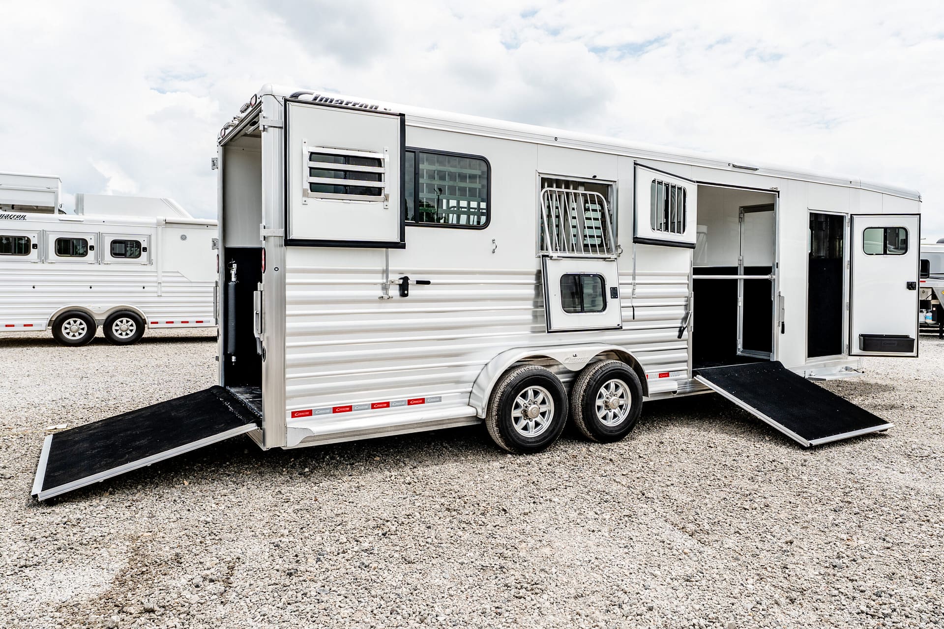 White horse trailer with open side ramps extended onto a gravel lot, other trailers visible in the background