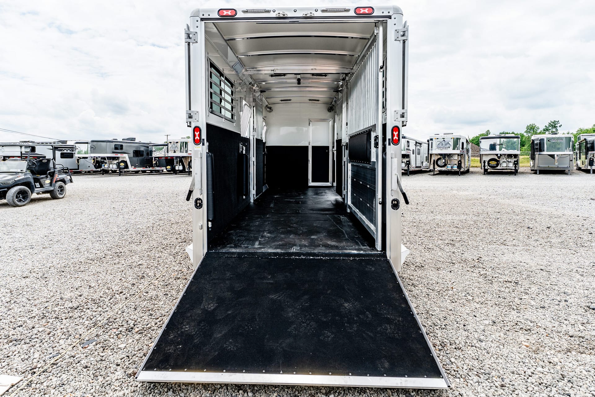 Open horse trailer with ramp lowered, ready for loading, in a gravel lot with other trailers in the background