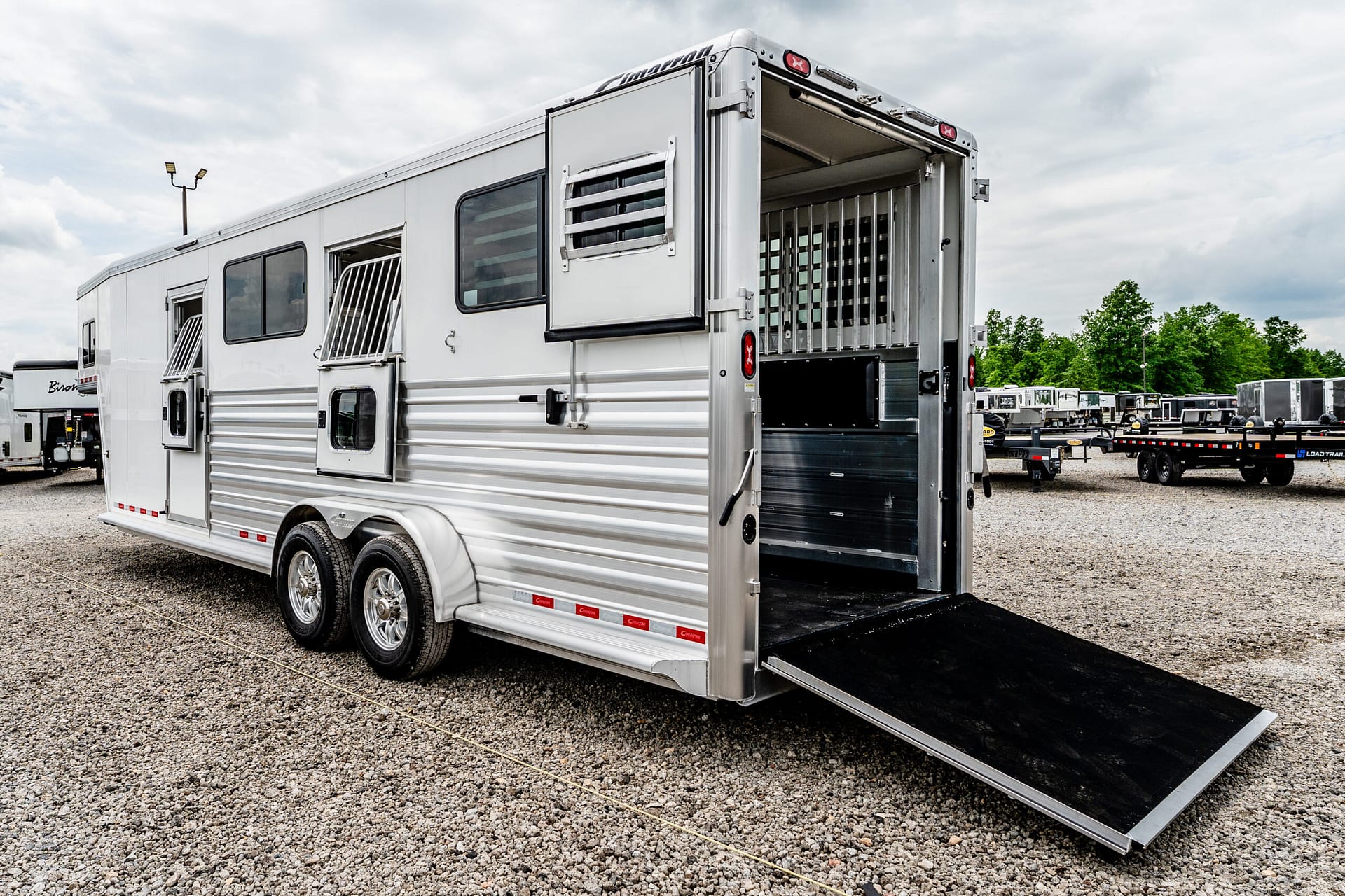 White horse trailer with open rear ramp in a gravel lot, multiple trailers visible in the background.