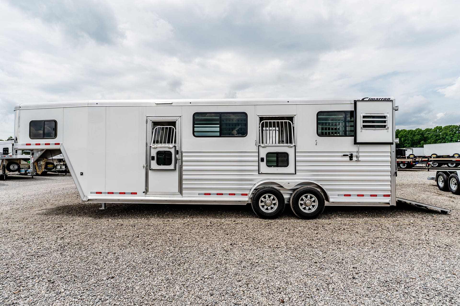White aluminum horse trailer on a gravel lot under a cloudy sky, with other trailers visible in the background.
