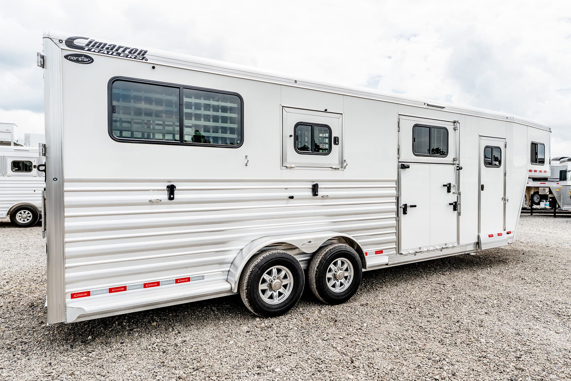 White horse trailer with windows and dual-axle wheels on a gravel lot, viewed from the side.