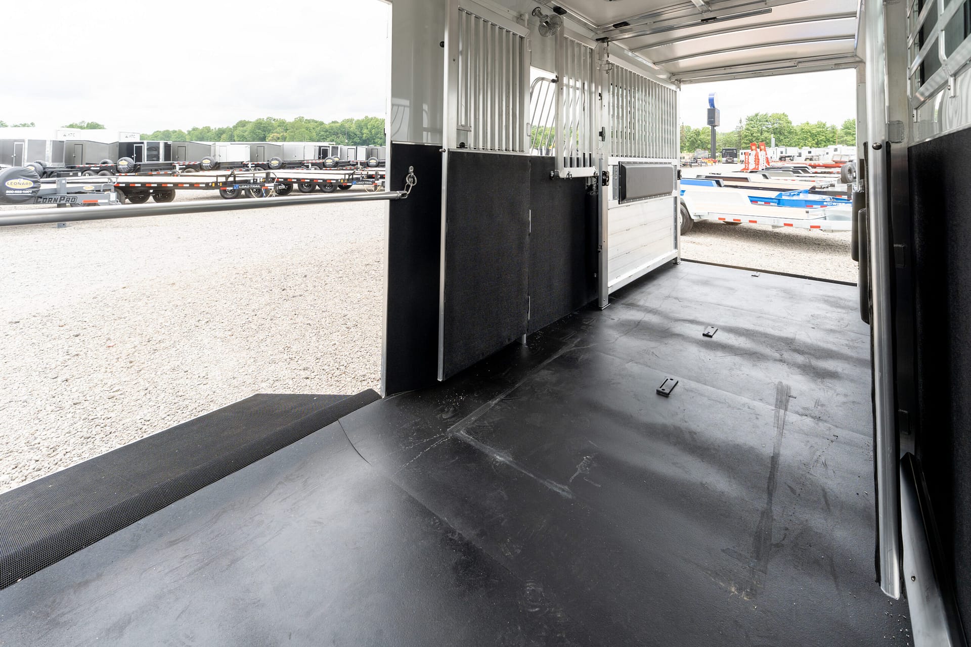 Interior of a large empty transport trailer with a black partition, metal walls, and an open ramp; outside is a gravel lot with many trailers parked.