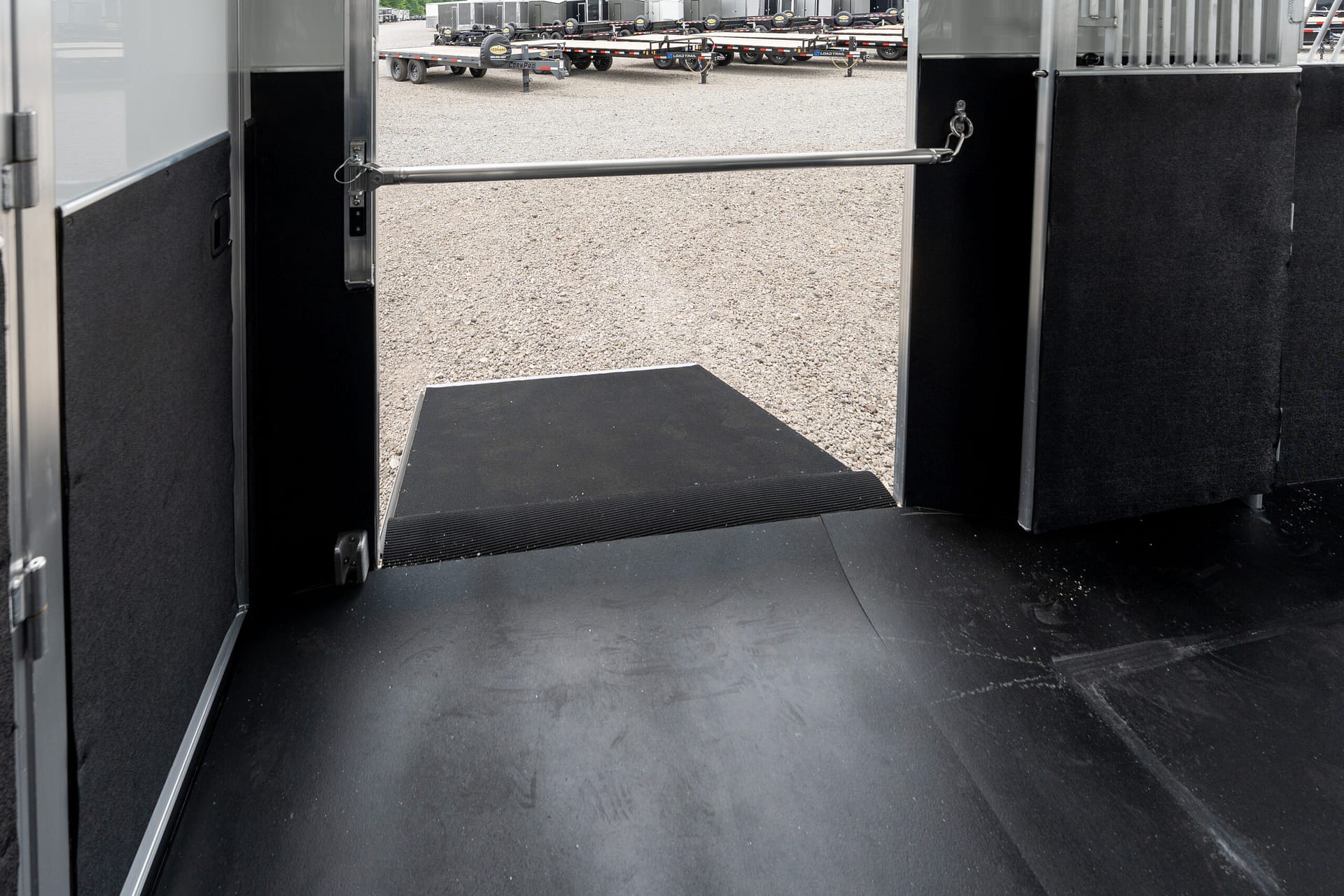 View from inside an open trailer with a lowered ramp, black interior walls, and a gravel yard with parked trailers in the distance.