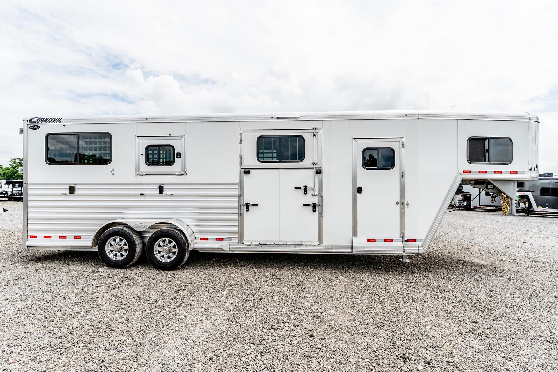 Long white horse trailer with multiple windows and doors on a gravel lot under a cloudy sky.