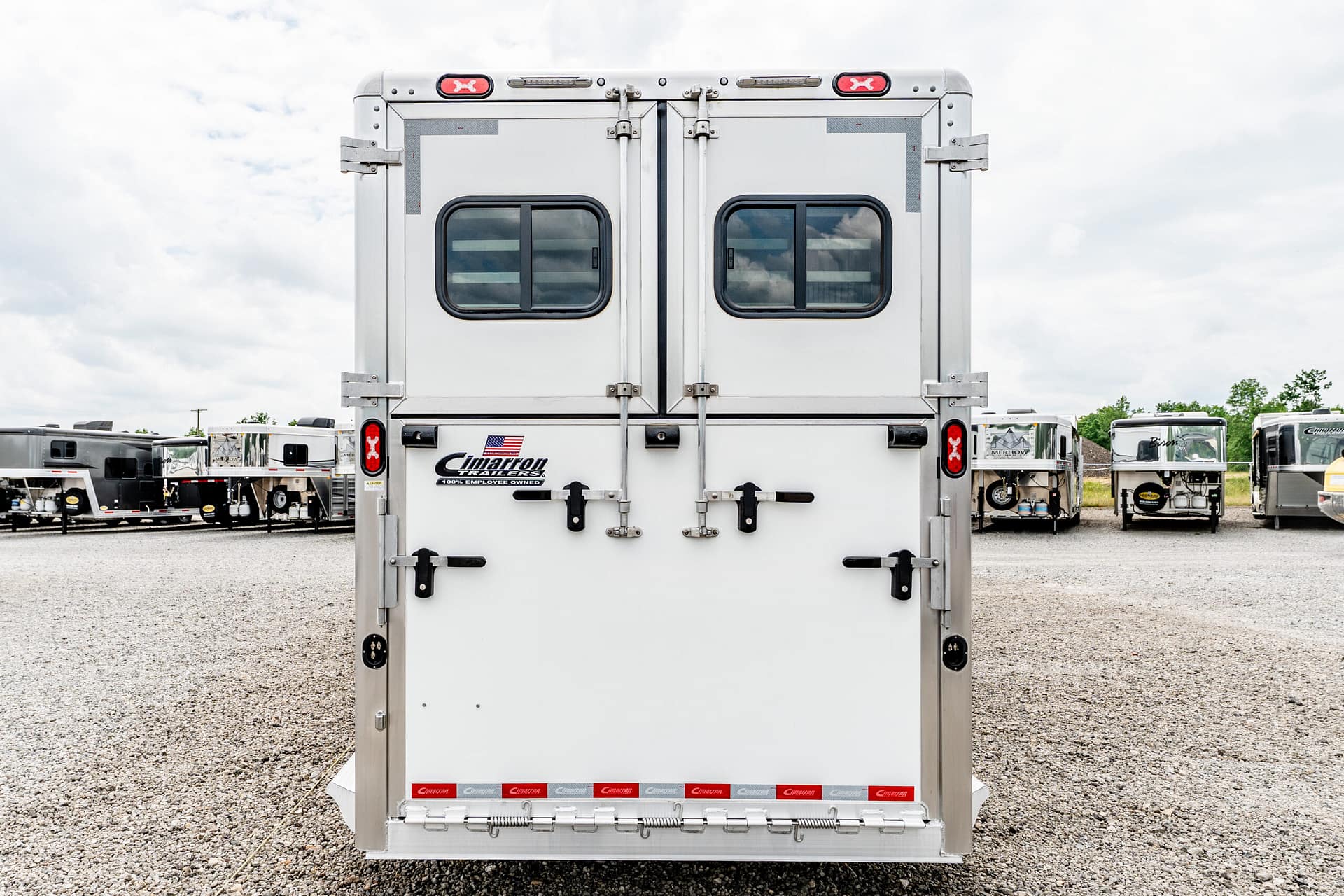 Rear view of a large aluminum cargo trailer with double doors and small windows, in a gravel lot with other trailers in the background.