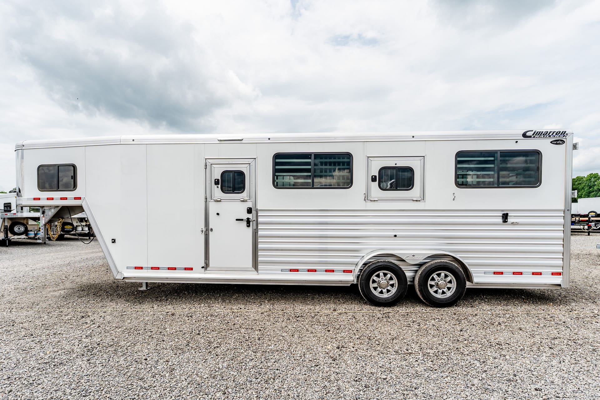 Long white horse trailer on a gravel lot under a cloudy sky, with doors and windows visible along the side.