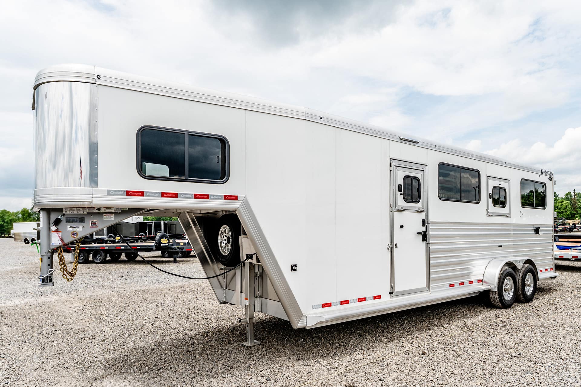 White horse trailer with a lowered ramp on a gravel lot beneath a cloudy sky, chrome trim along the sides.