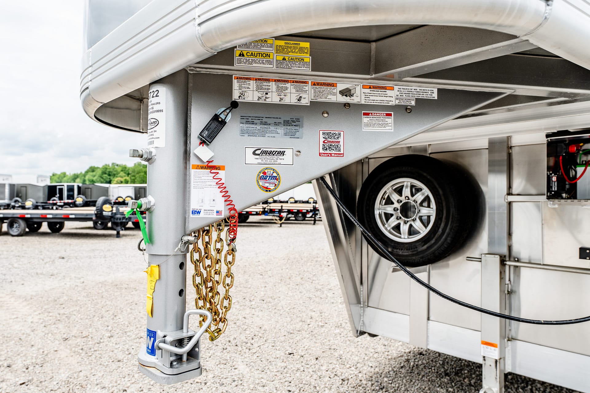 Close-up of a trailer hitch area with safety chains and warning stickers on a silver trailer frame, wheel in background, outdoors