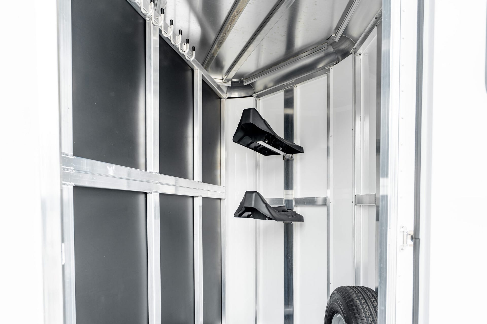 Interior of a metal trailer showing wall-mounted hooks, framing, and a black wheel chock along a vertical track with a spare tire at the bottom left.
