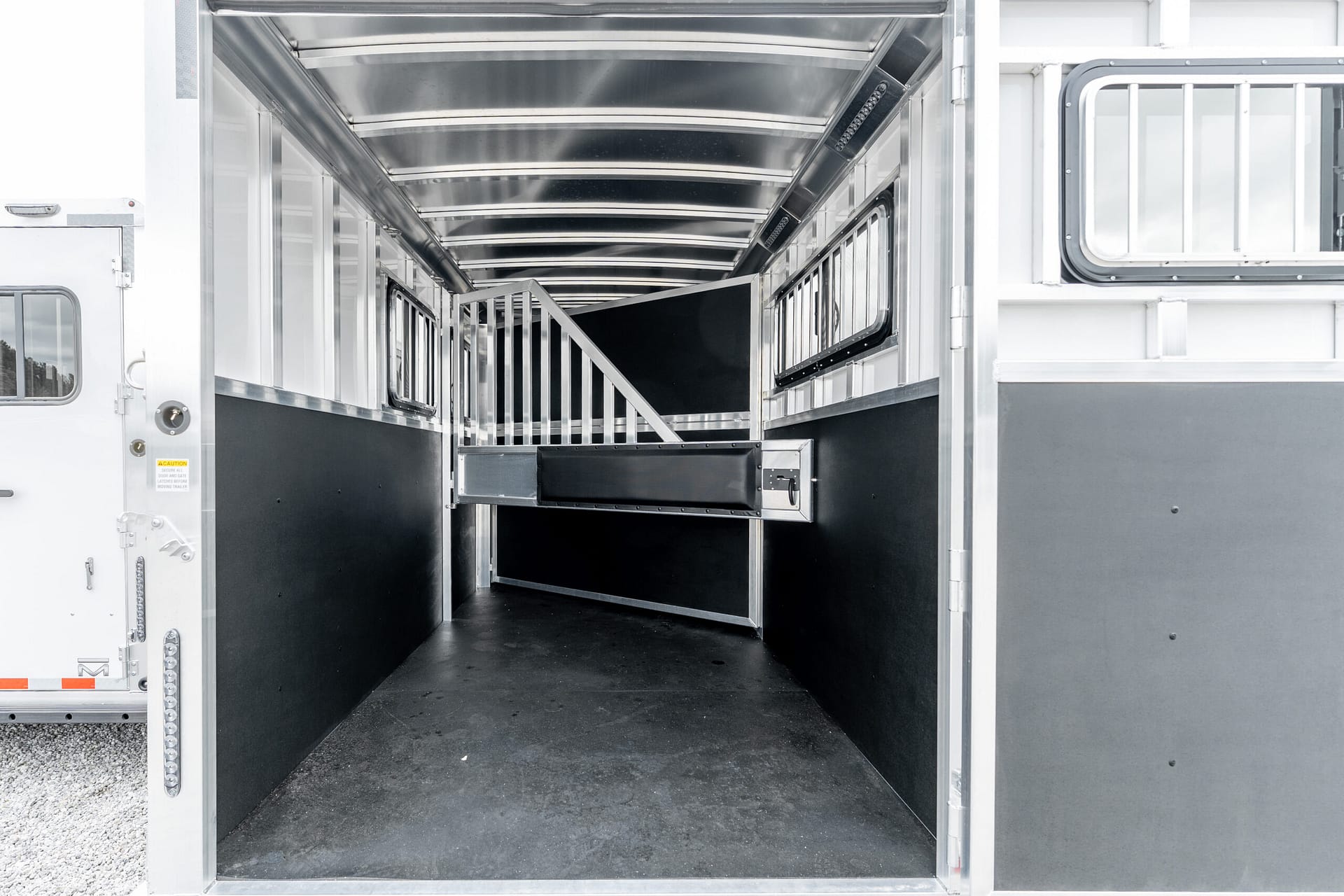 Interior of a silver horse trailer with metal stalls and dividing rails, leading to a ramp at the far end; bright, industrial inside.