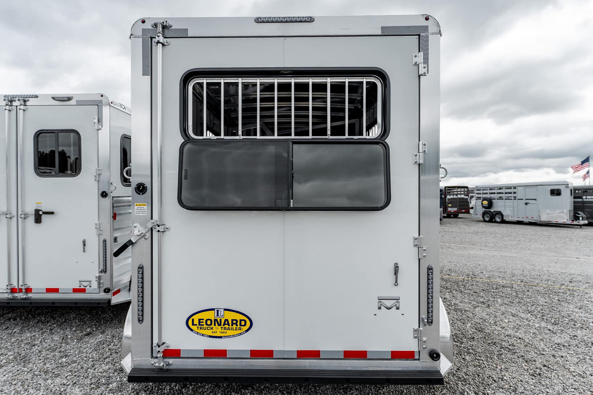 Rear view of a silver cargo trailer with an open ventilated window and vertical bars, Leonard Truck & Trailer logo on the back, parked on gravel with other trailers nearby.