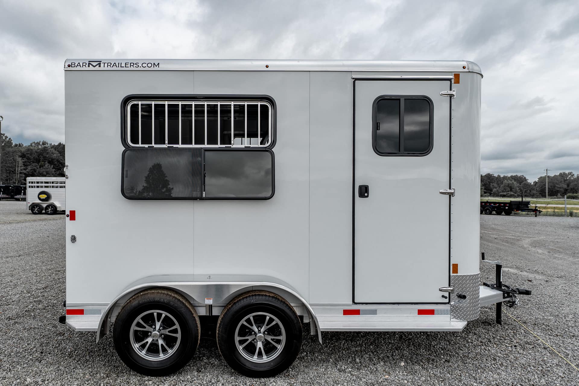 Side view of a silver horse trailer with barred window and dual rear wheels on a gravel lot
