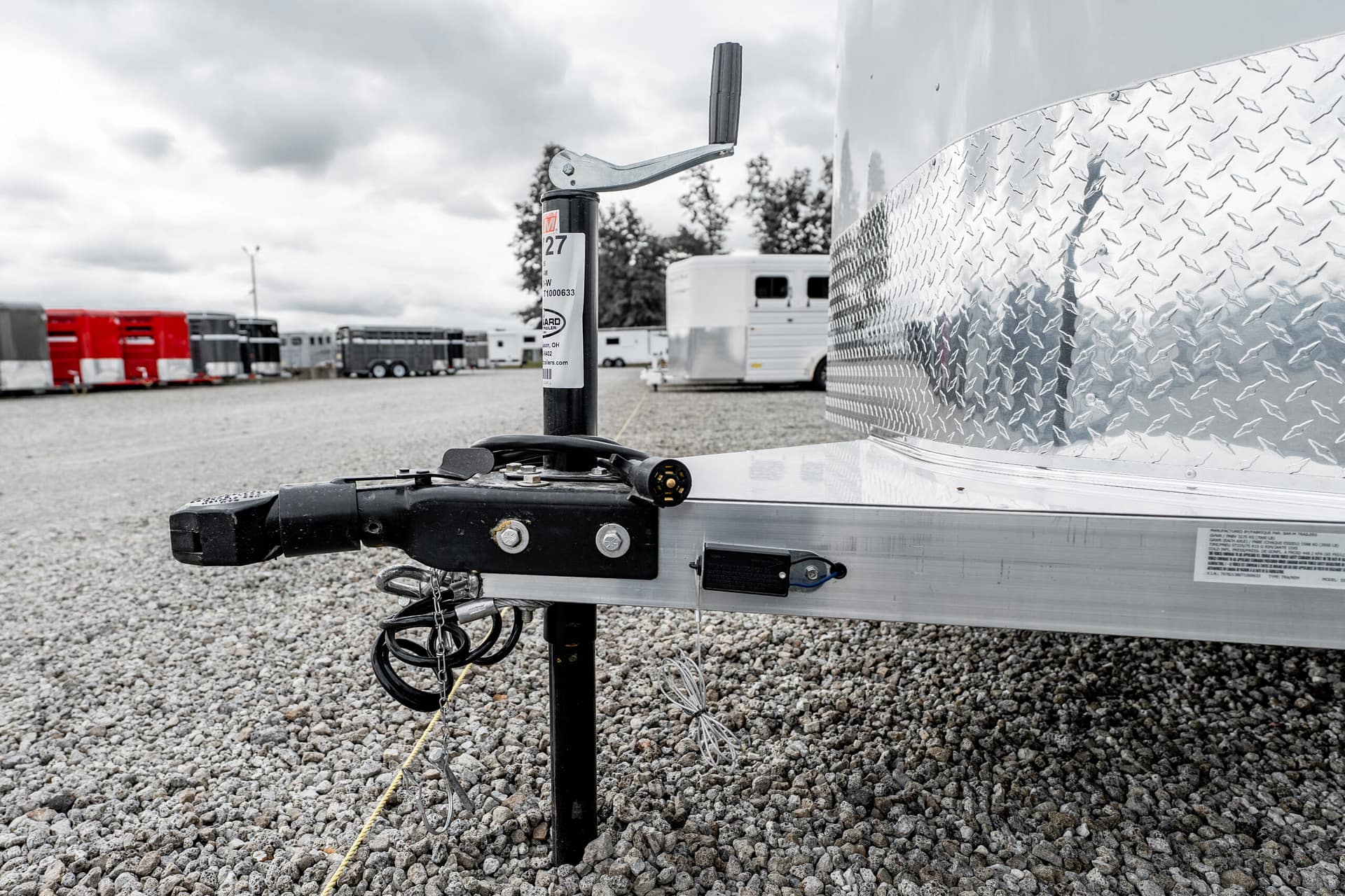 Close-up view of a trailer hitch and jack on a gravel lot, with diamond-plate trailer side and other trailers in the background.