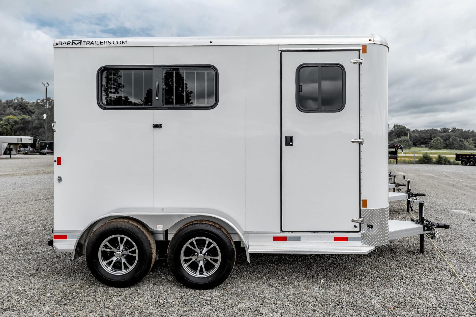 White utility trailer with two axles and a large side window, parked on a gravel lot under a cloudy sky
