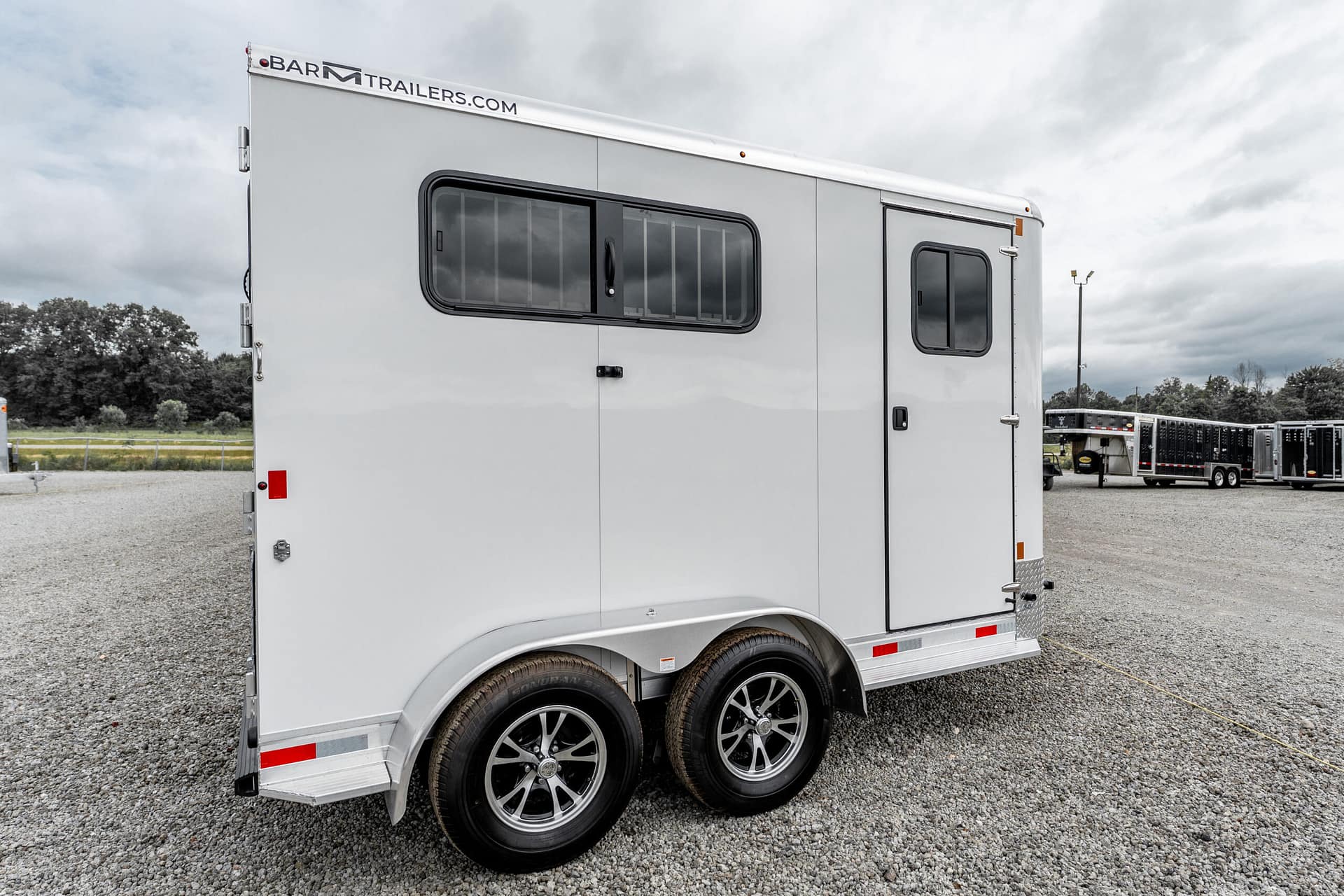 Long silver horse trailer with two axles and a rounded, tinted window, parked on a gravel lot with other trailers in the background and a BARN TRAILERS banner along the top edge.