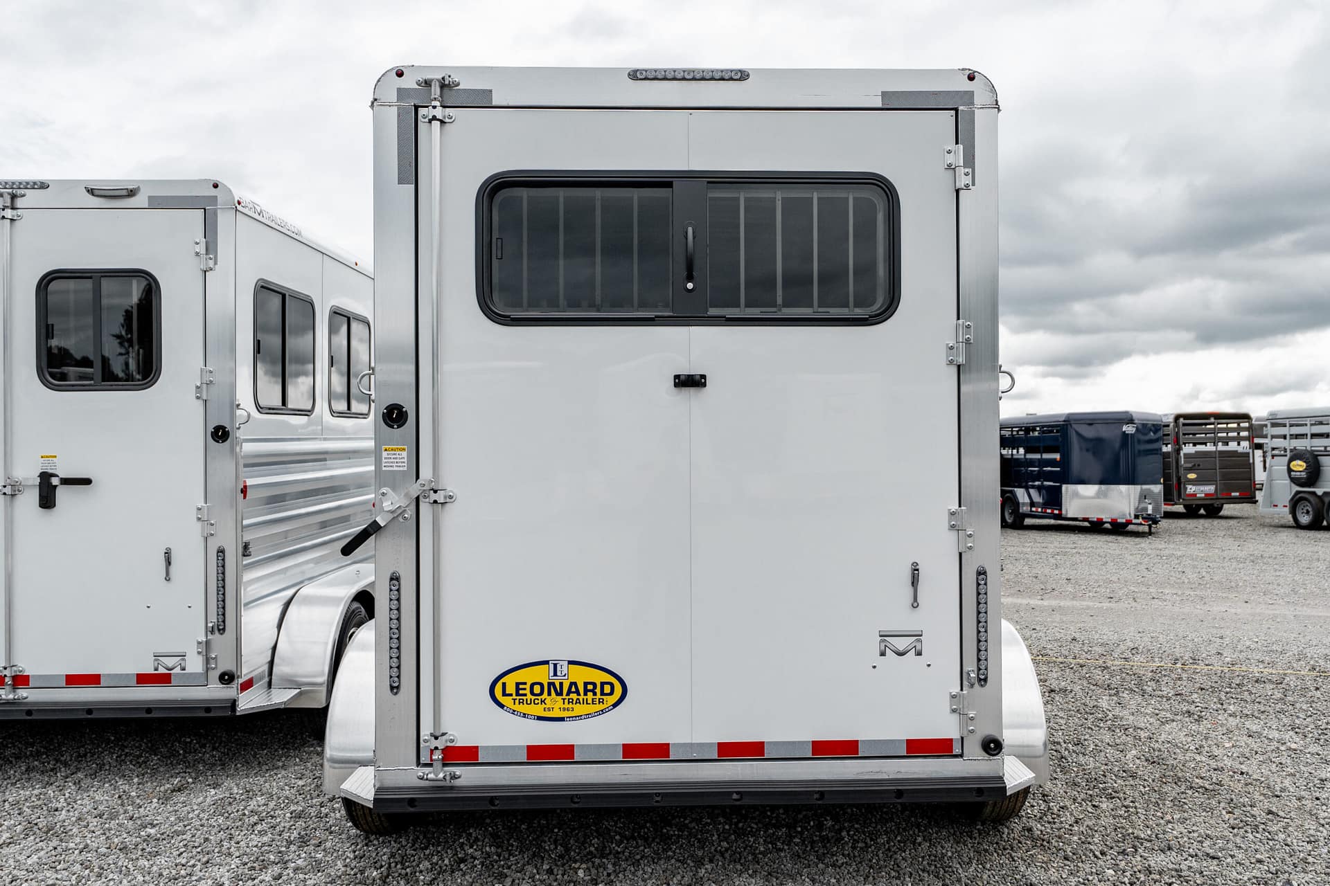 Rear view of a silver semi-trailer with barred rear window and Leonard Truck & Trailer logo on the lower back, parked on gravel.