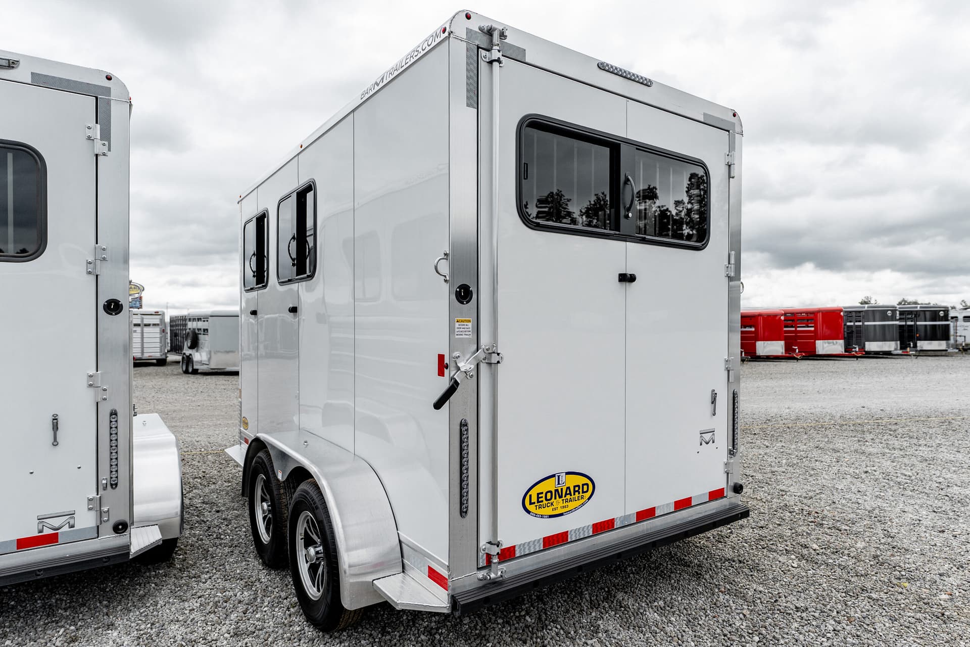 Rear view of a silver trailer with Leonard Truck & Trailer logo on a gravel lot under a cloudy sky, with red-and-white reflectors visible.