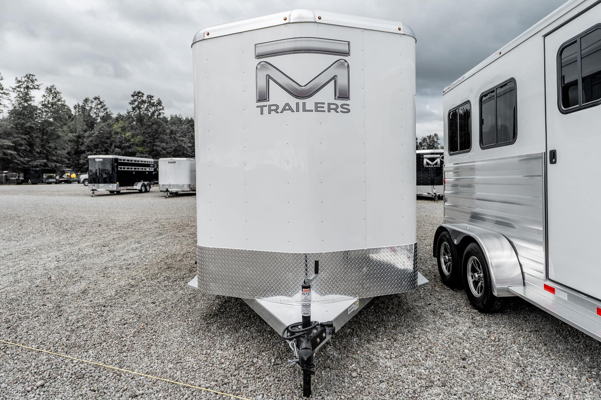 Close-up of a white cargo trailer tongue and hitch on a gravel lot, with other trailers and trees in the background.