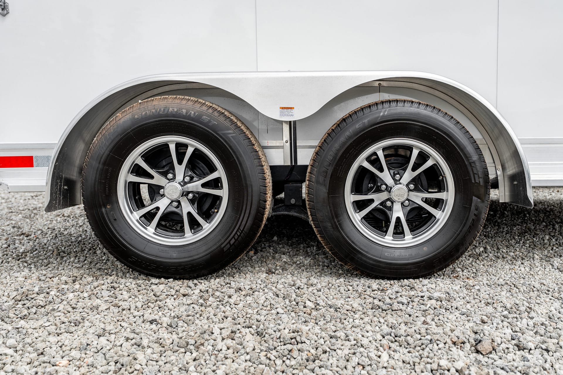 Dual trailer wheels with silver rims under a curved aluminum fender on a gravel surface.