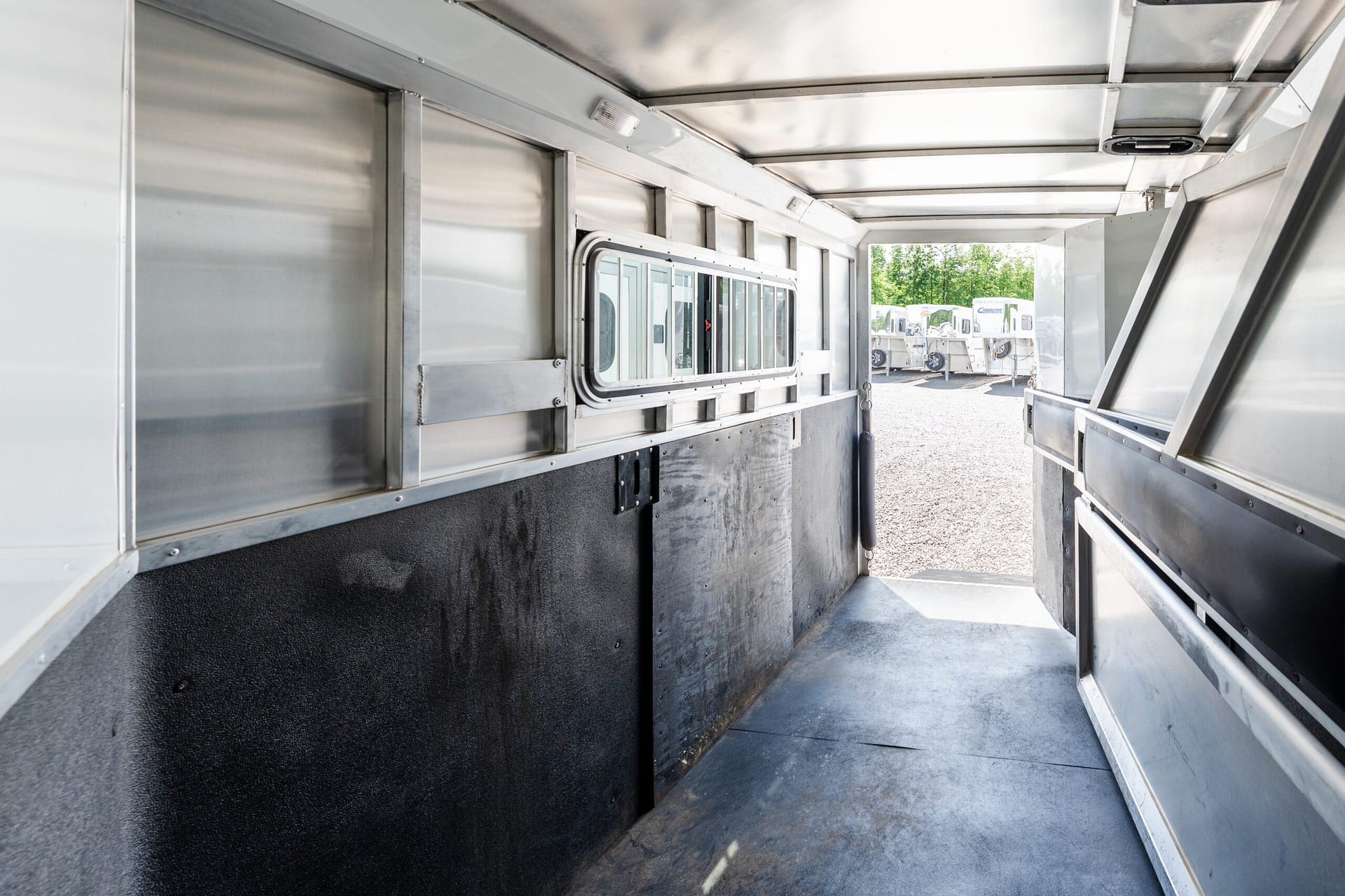 Interior of a silver metal trailer with a row of windows along the left wall and an open rear doorway showing a gravel lot and other trailers outside.