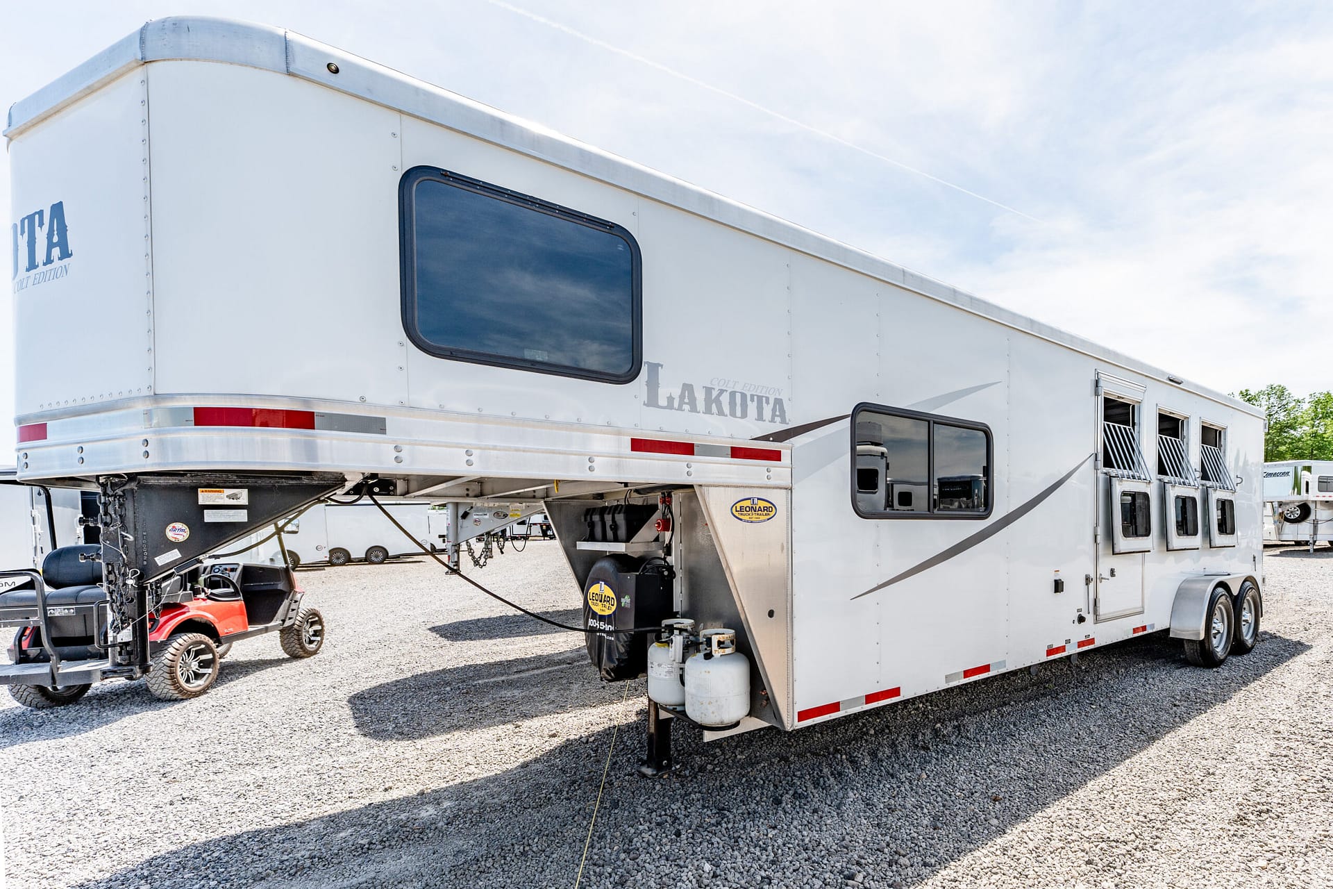 White Lakota horse trailer with a large rear-side window, parked on a gravel lot under a clear blue sky.