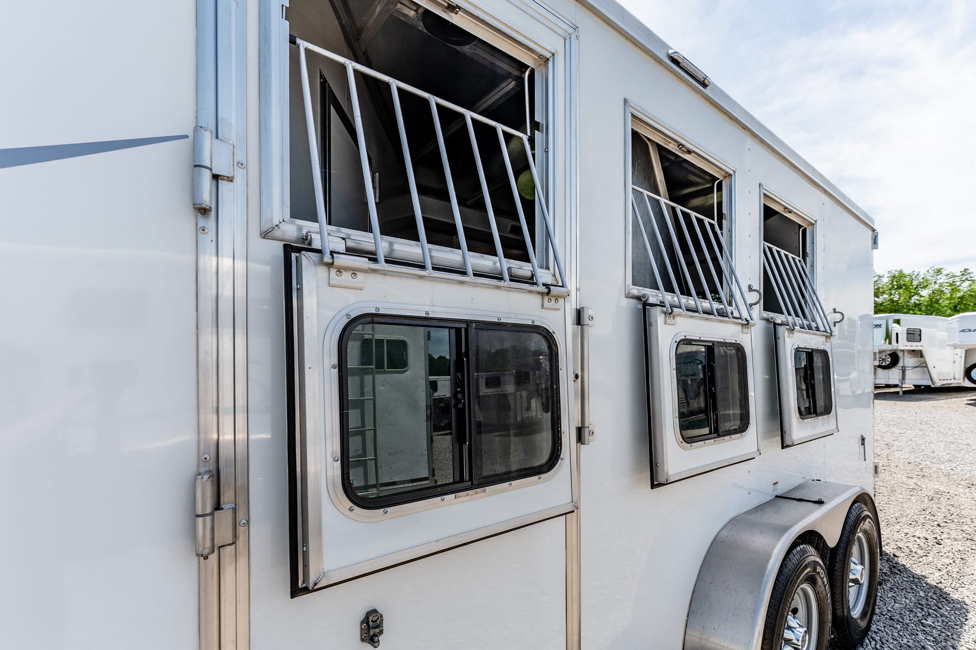 Side view of a white horse trailer with metal-barred windows and chrome trim, parked on a gravel lot.