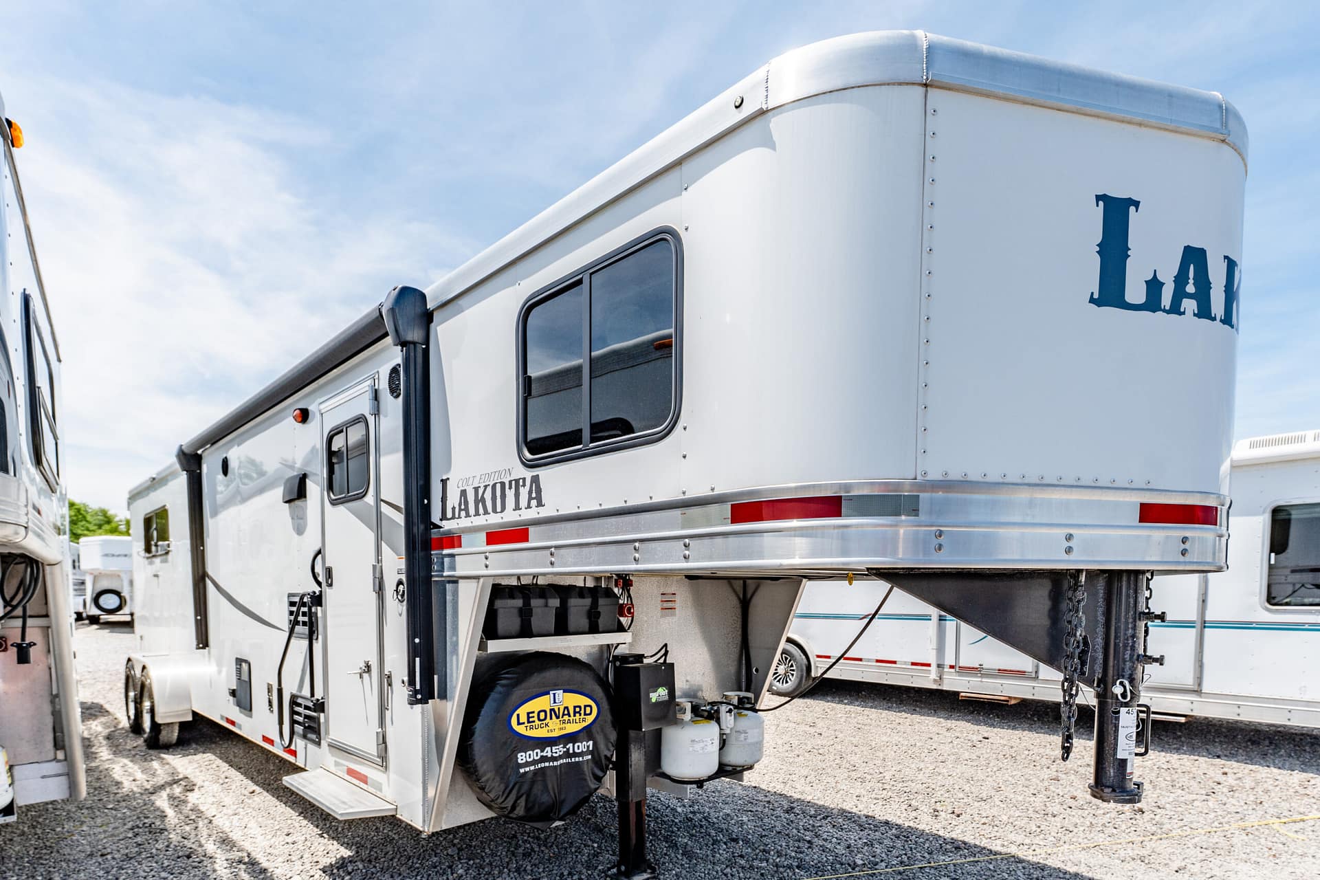 White Lakota horse trailer on a gravel lot, showing a side window, entry door, and spare tire with Leonard branding.